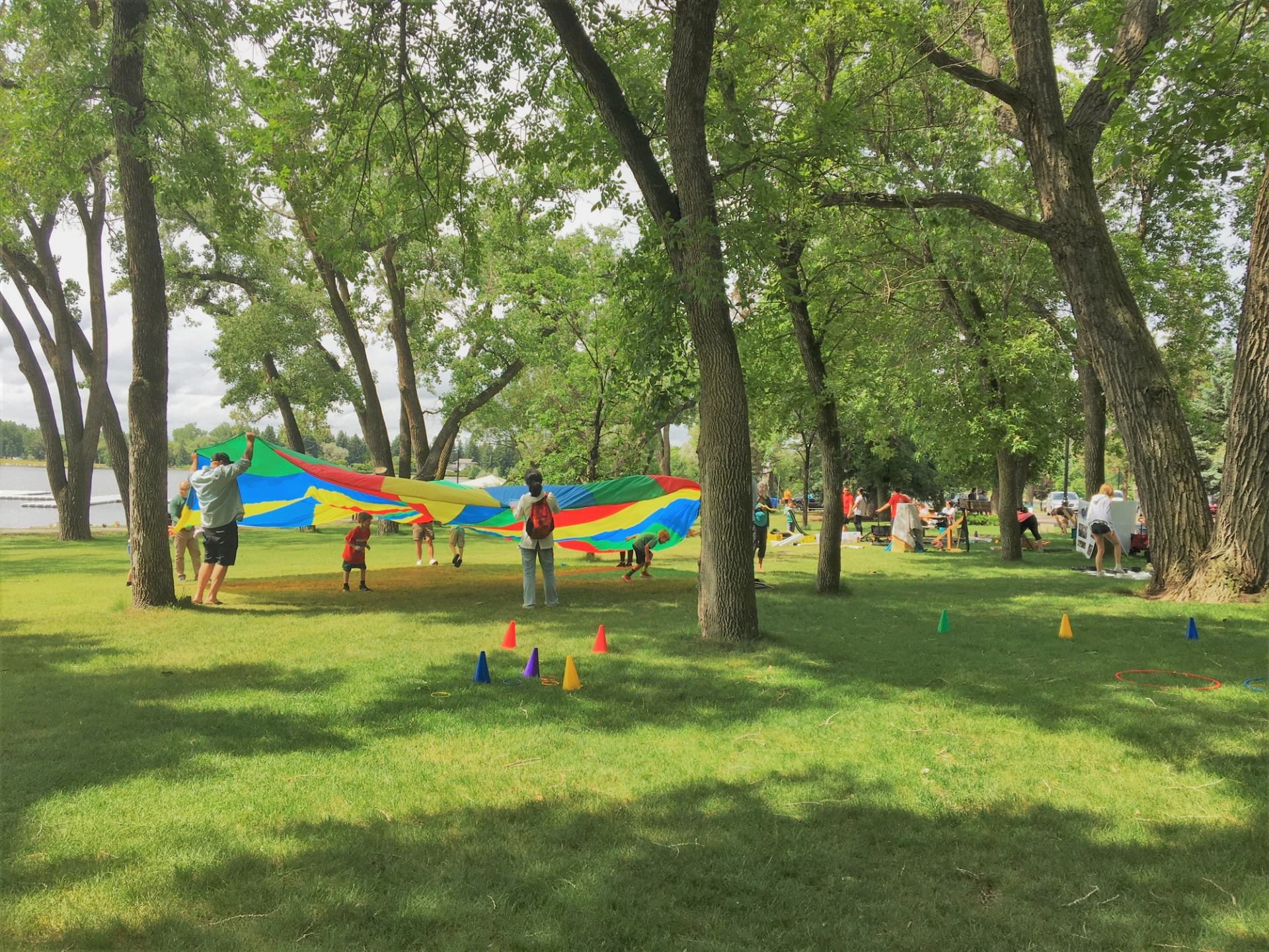 Children and adults play with a large rainbow parachute in a grassy, tree-filled park.