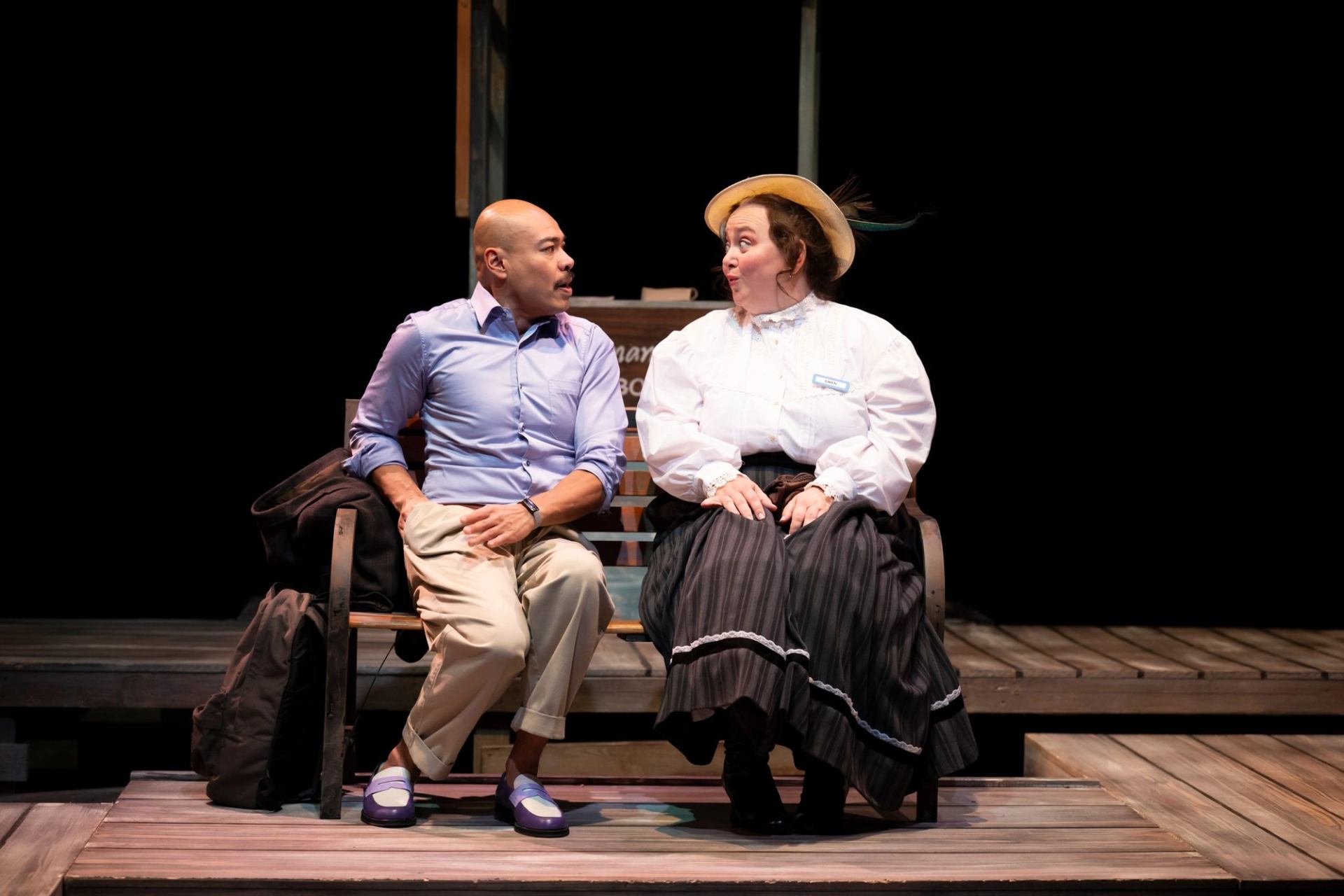 Stage actors sit on a wooden bench, talking during a theatre performance.