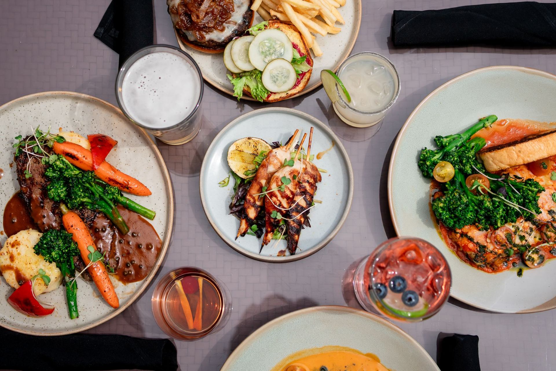 Chicken Skewers surrounded by a steak dish, open faced burger and lasagna with drinks.