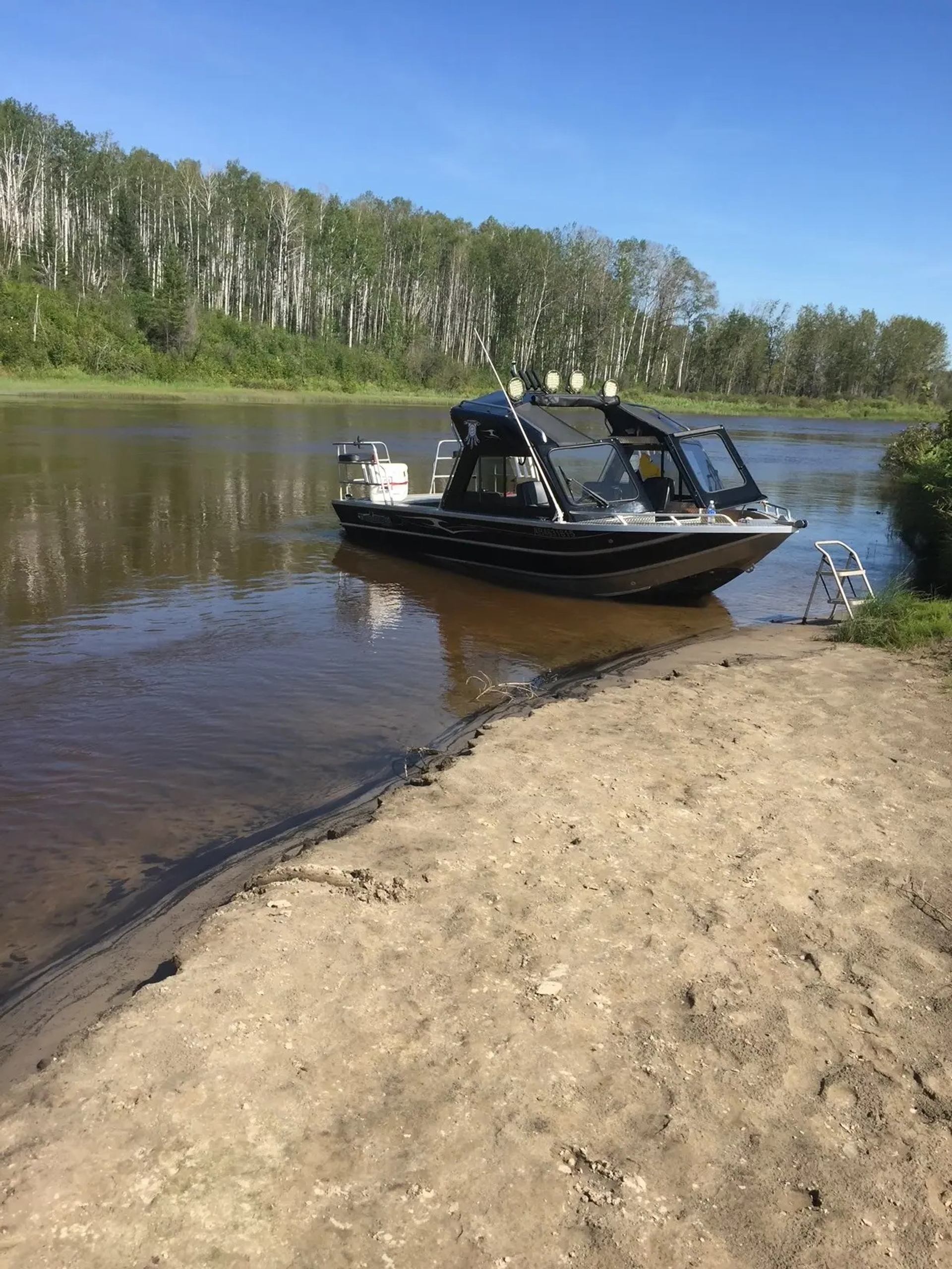 Aluminium motorboat moored on calm river beside sandy shore and forested bank.