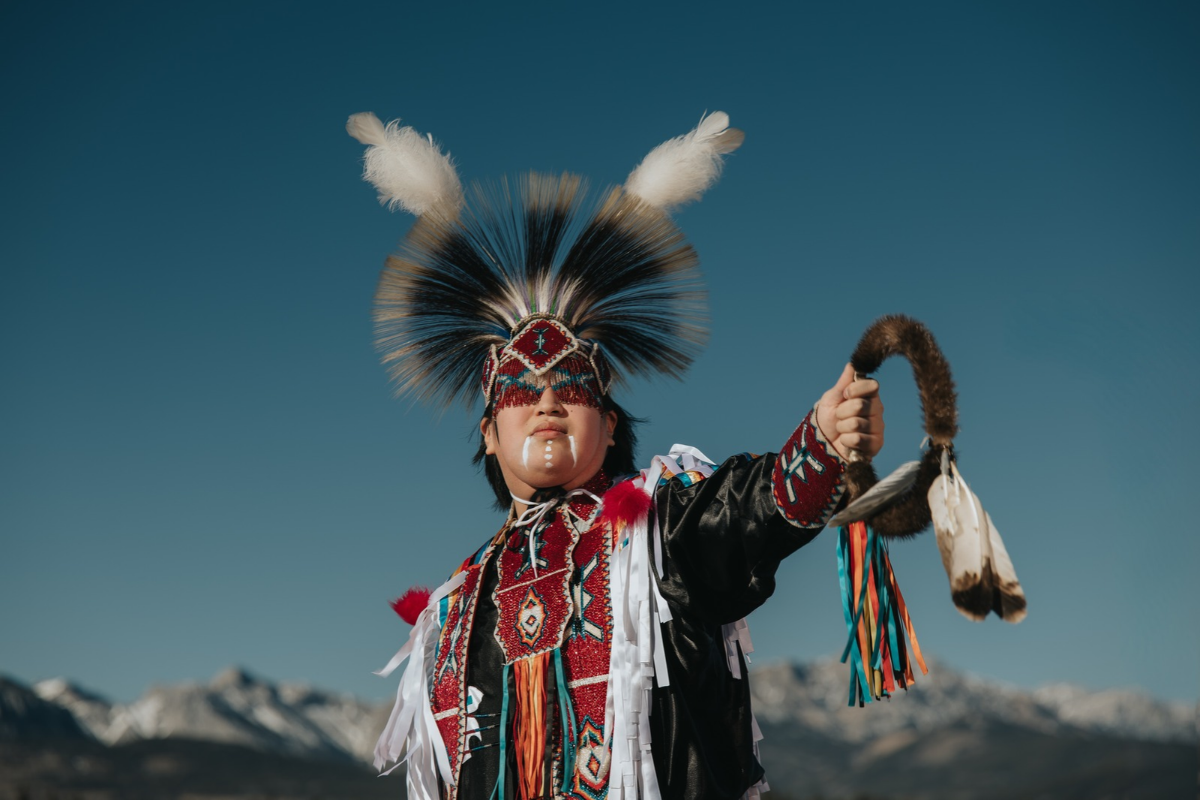 Person in feathered regalia holds a fur-wrapped hoop before distant mountains.