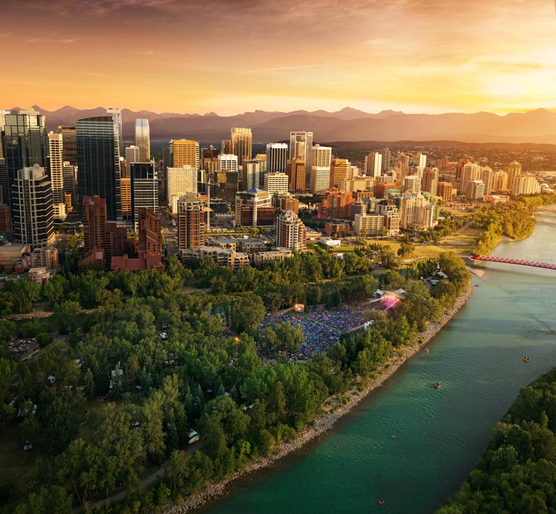 City skyline with tall buildings, riverside park, outdoor crowd and distant mountains.