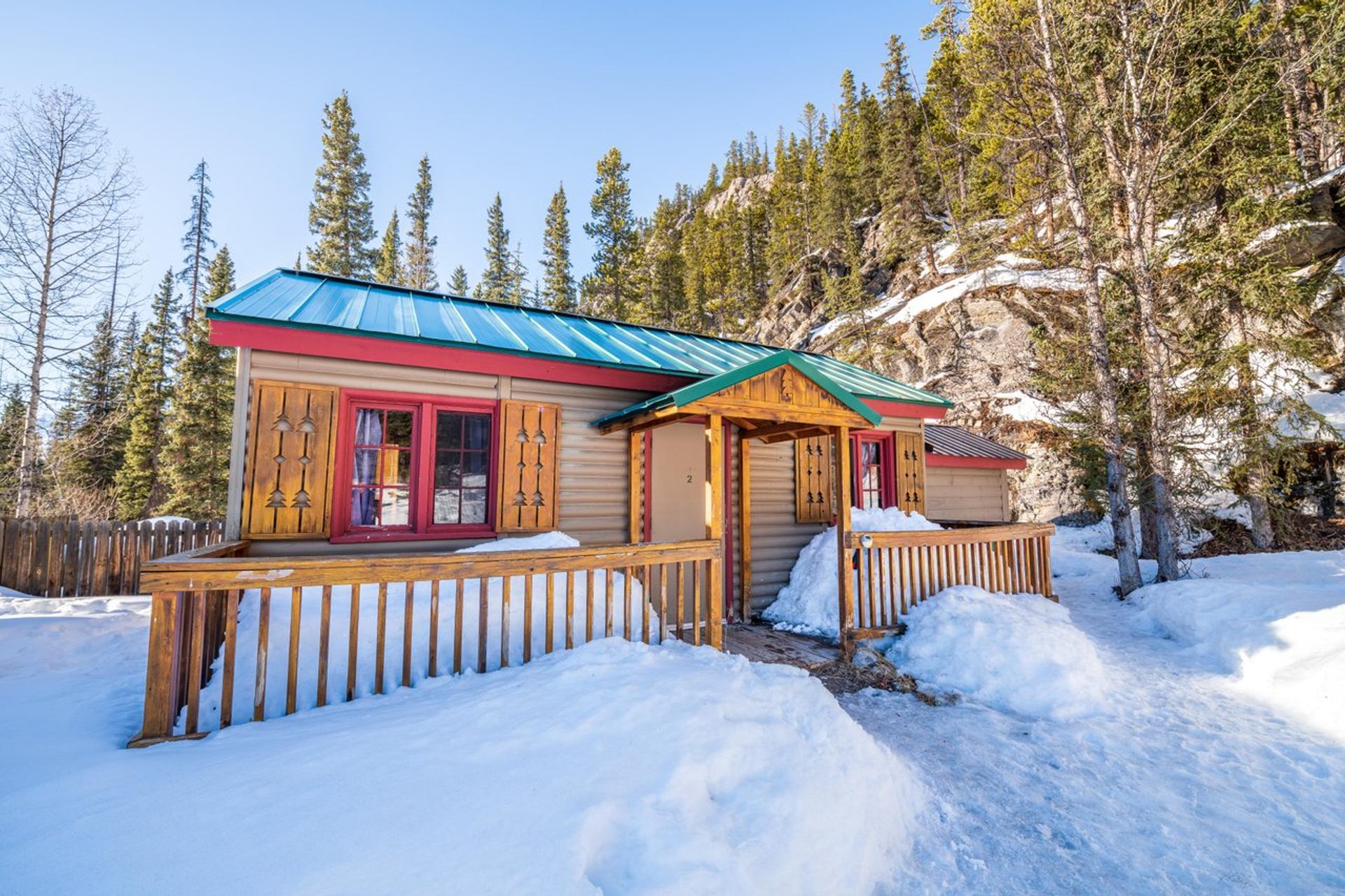 Snow-covered cabin with red trim, solar panels, and forested mountain backdrop.