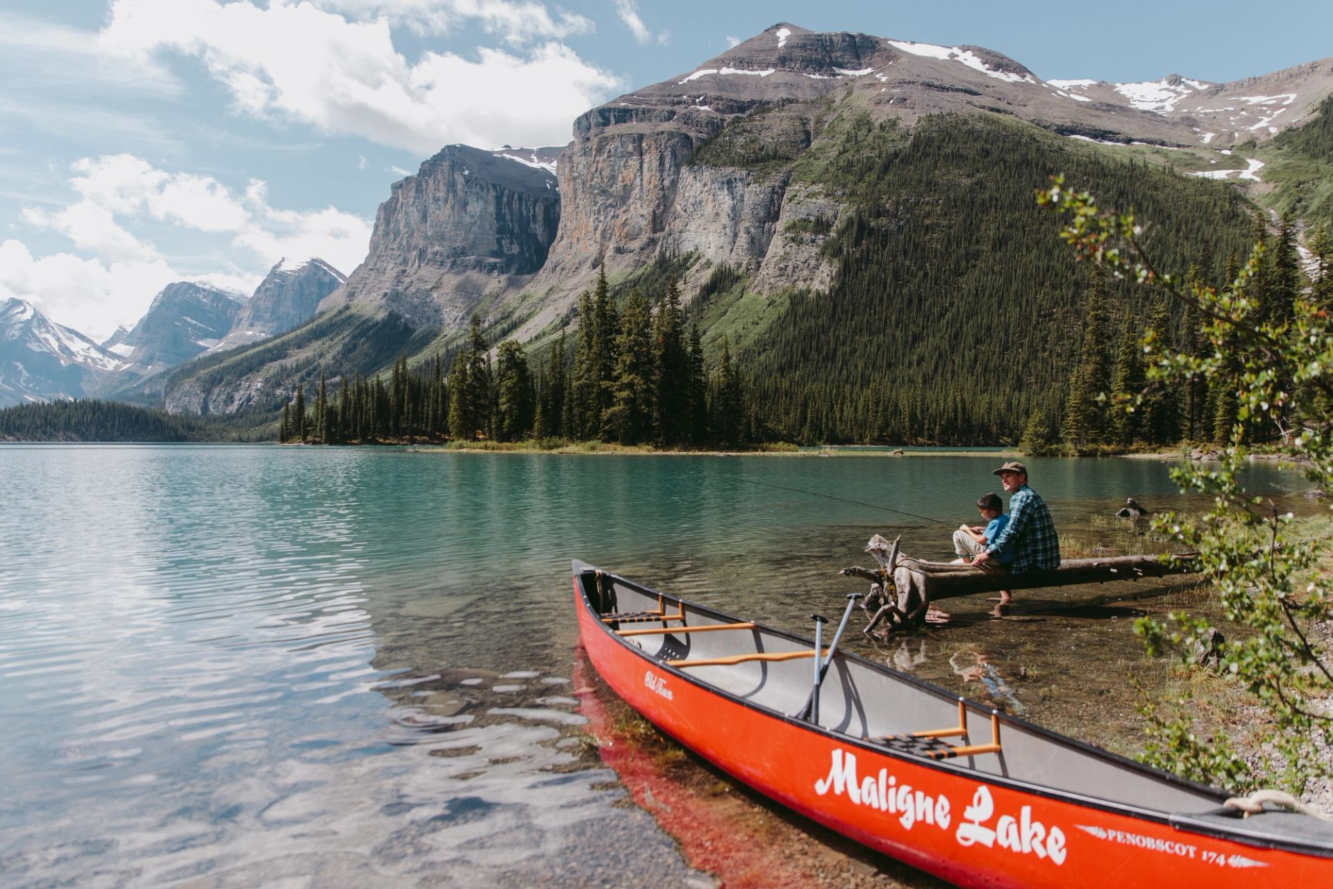 Man and child sitting on a log with a red canoe on Maligne Lake.