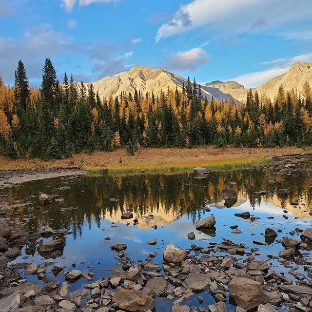 A serene mountain scene with a still lake reflecting rocky peaks and autumnal trees. The sky is blue with some clouds, conveying a tranquil atmosphere.