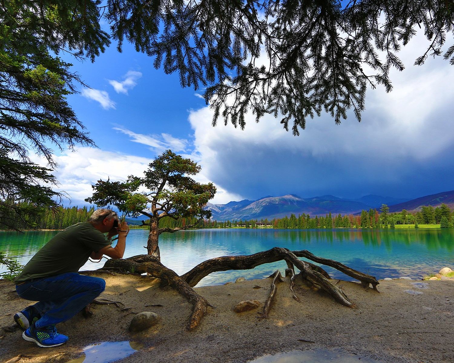 A person photographing the scenic landscape with a lake, mountains, and trees in the background.
