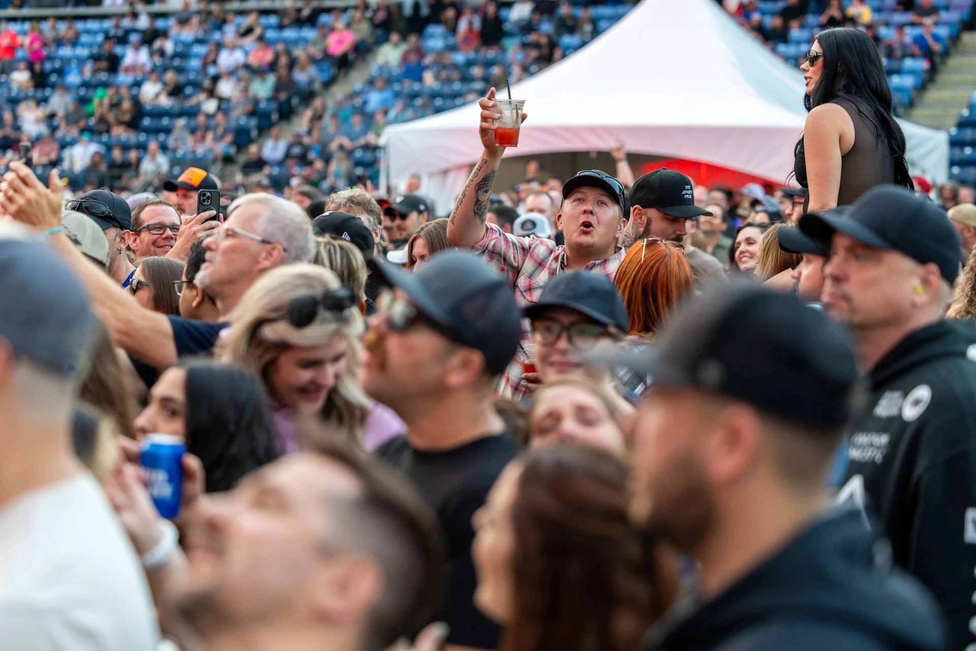 Concert crowd in stadium, with person raising drink cup and others watching the stage.