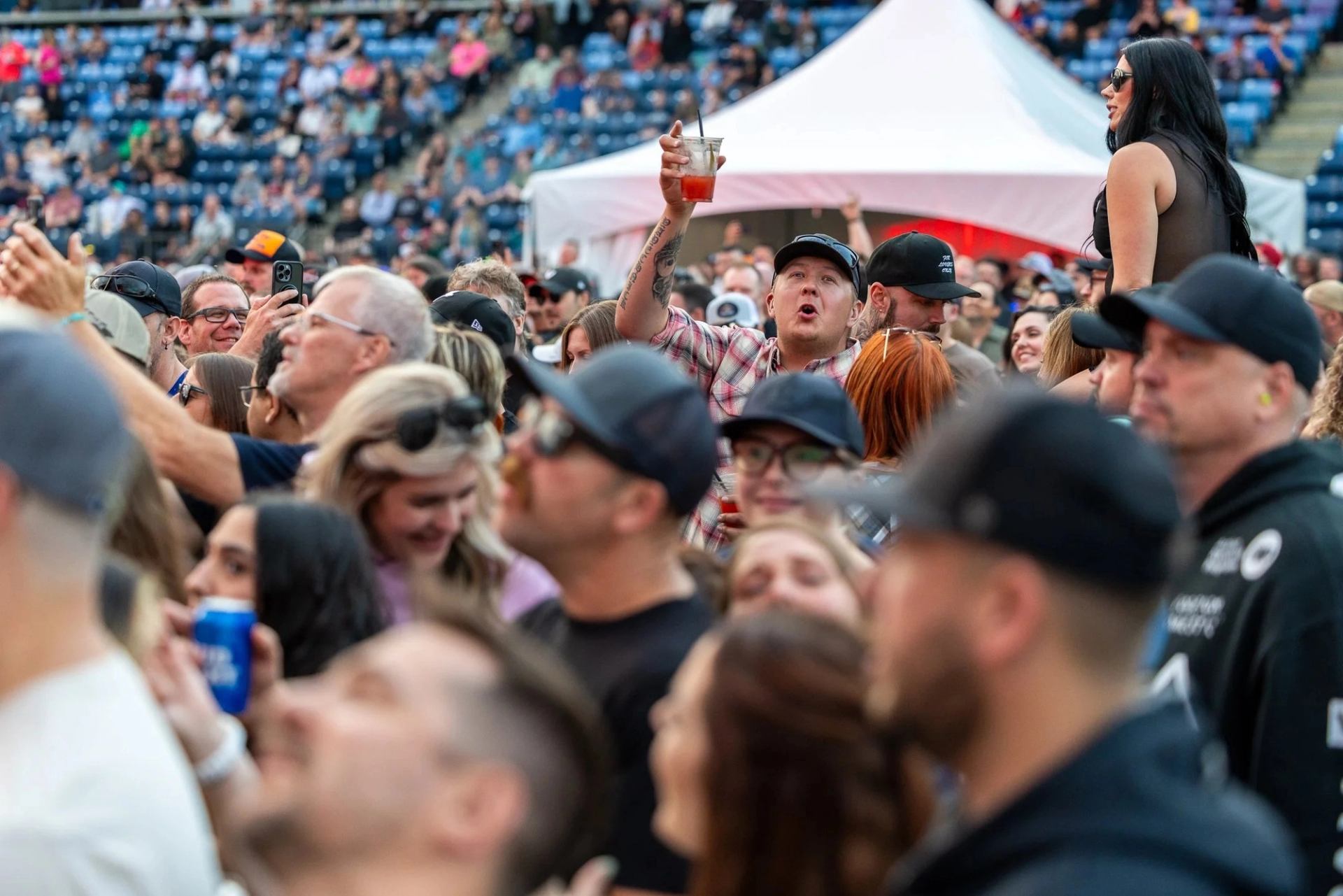 Concert crowd in stadium, with person raising drink cup and others watching the stage.