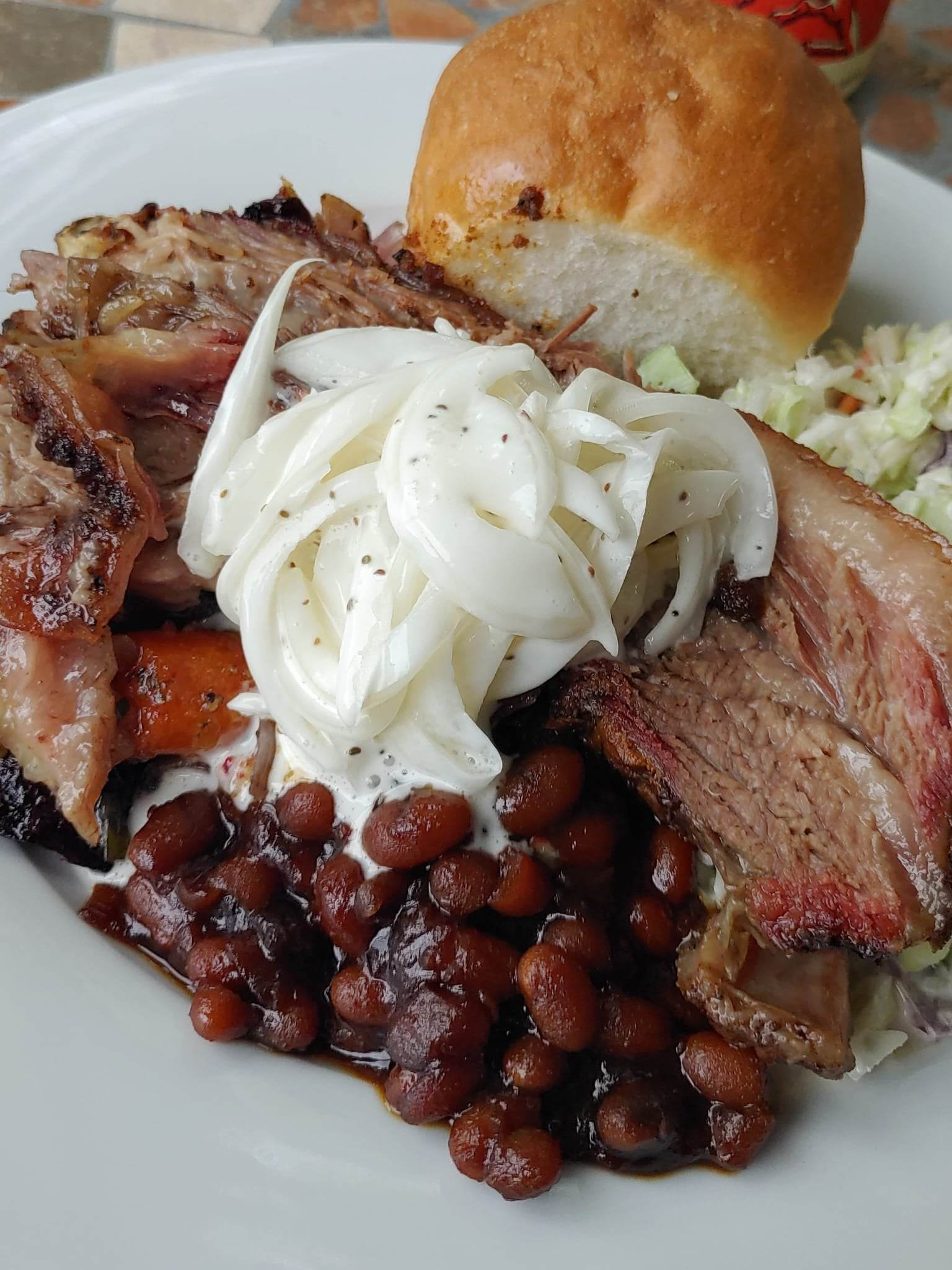 Plate of barbecued meat with baked beans, creamy onions, coleslaw and a bun.