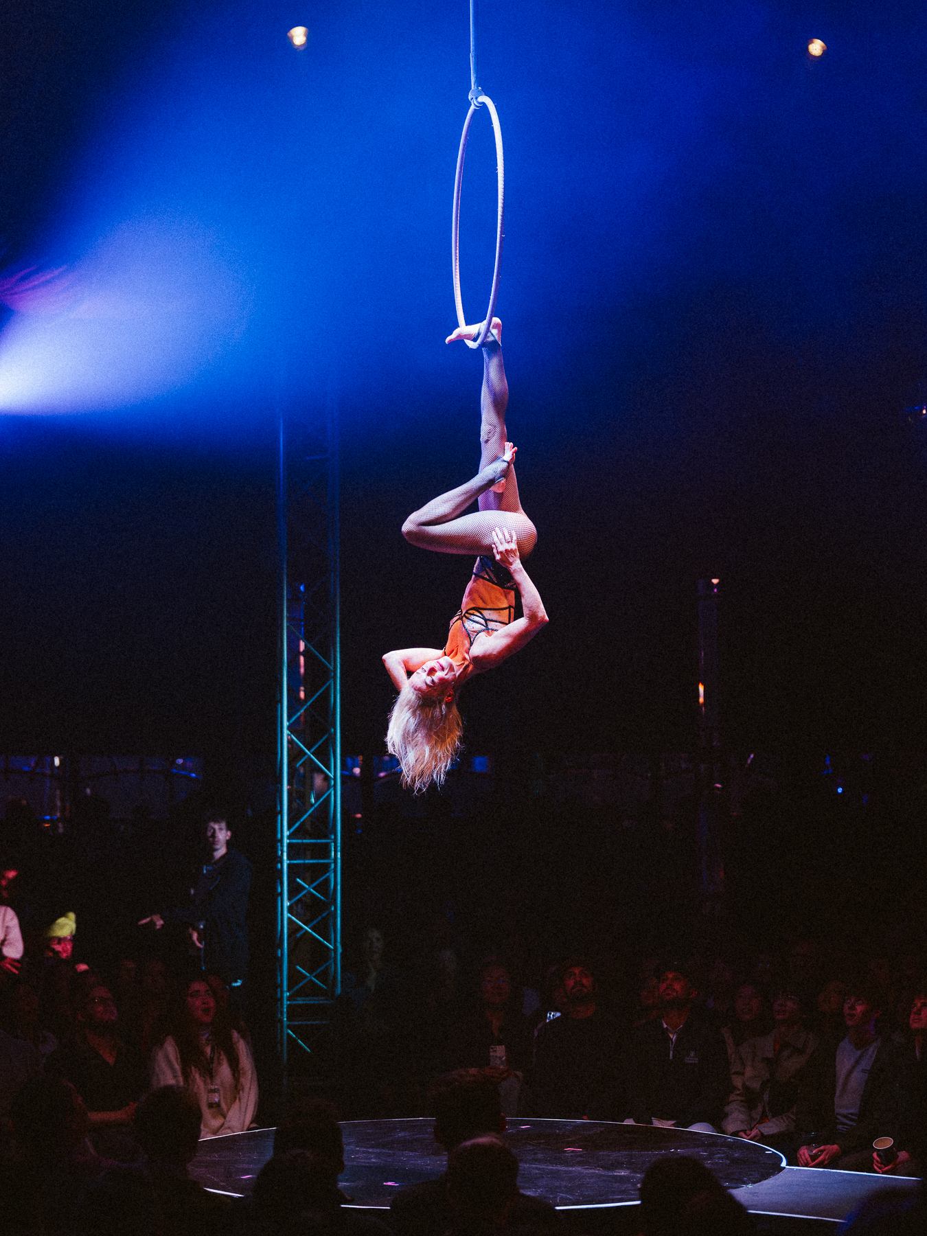 Aerial performer hangs upside down from hoop above stage, audience watching below.