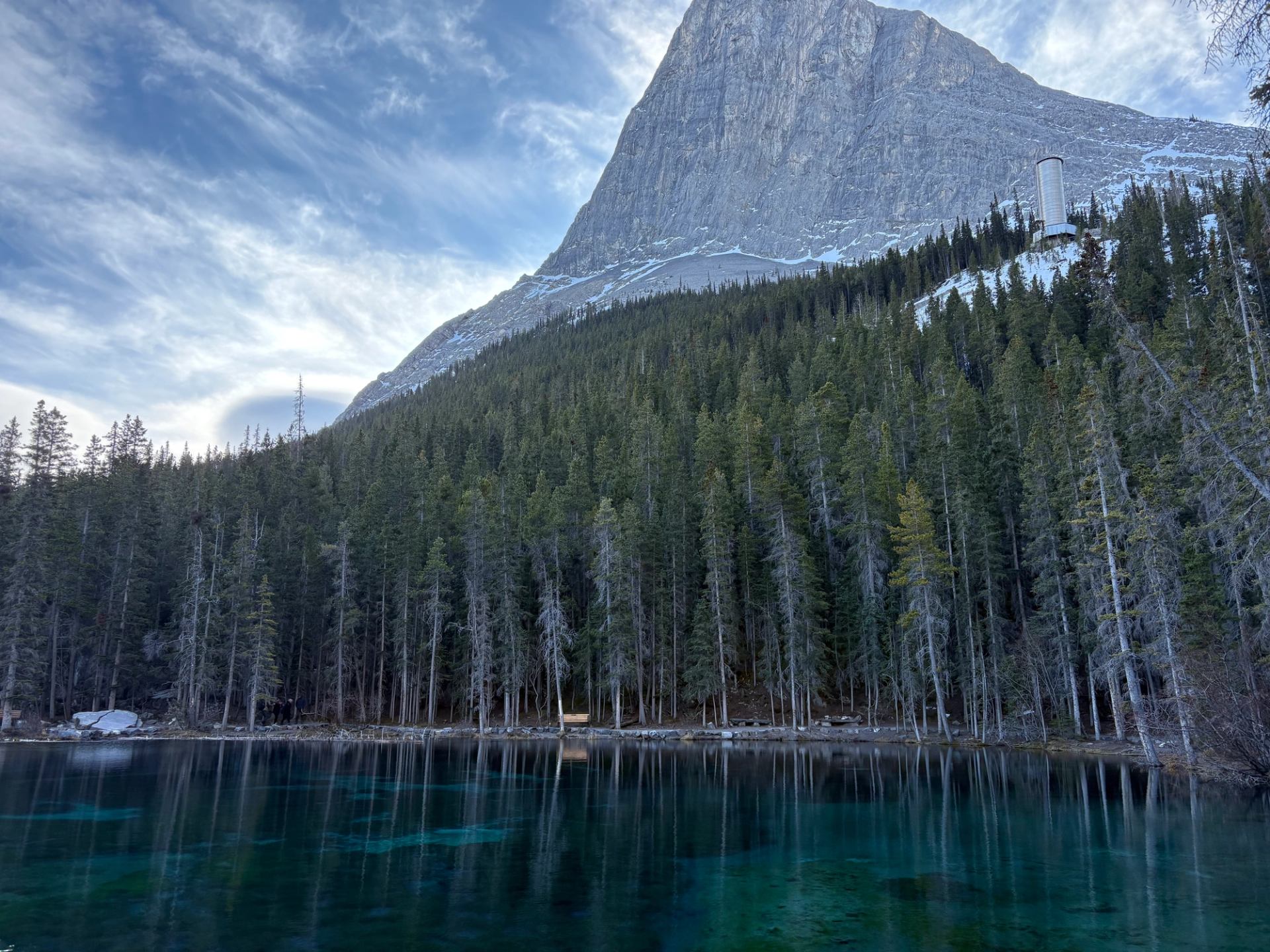 Mountain towering above dense evergreen forest reflected in clear turquoise lake under wispy clouds.