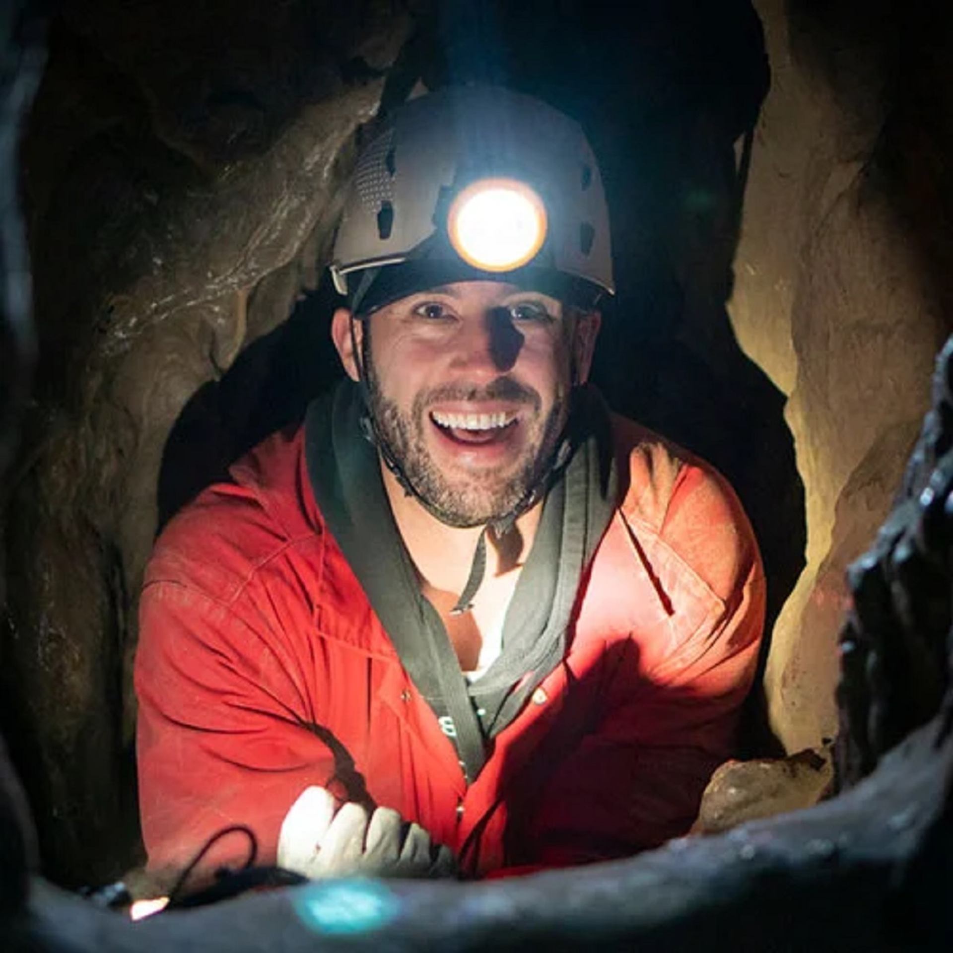 Helmeted caver in a red jacket explores a narrow limestone passage inside Rat’s Nest Cave, lit by a headlamp.