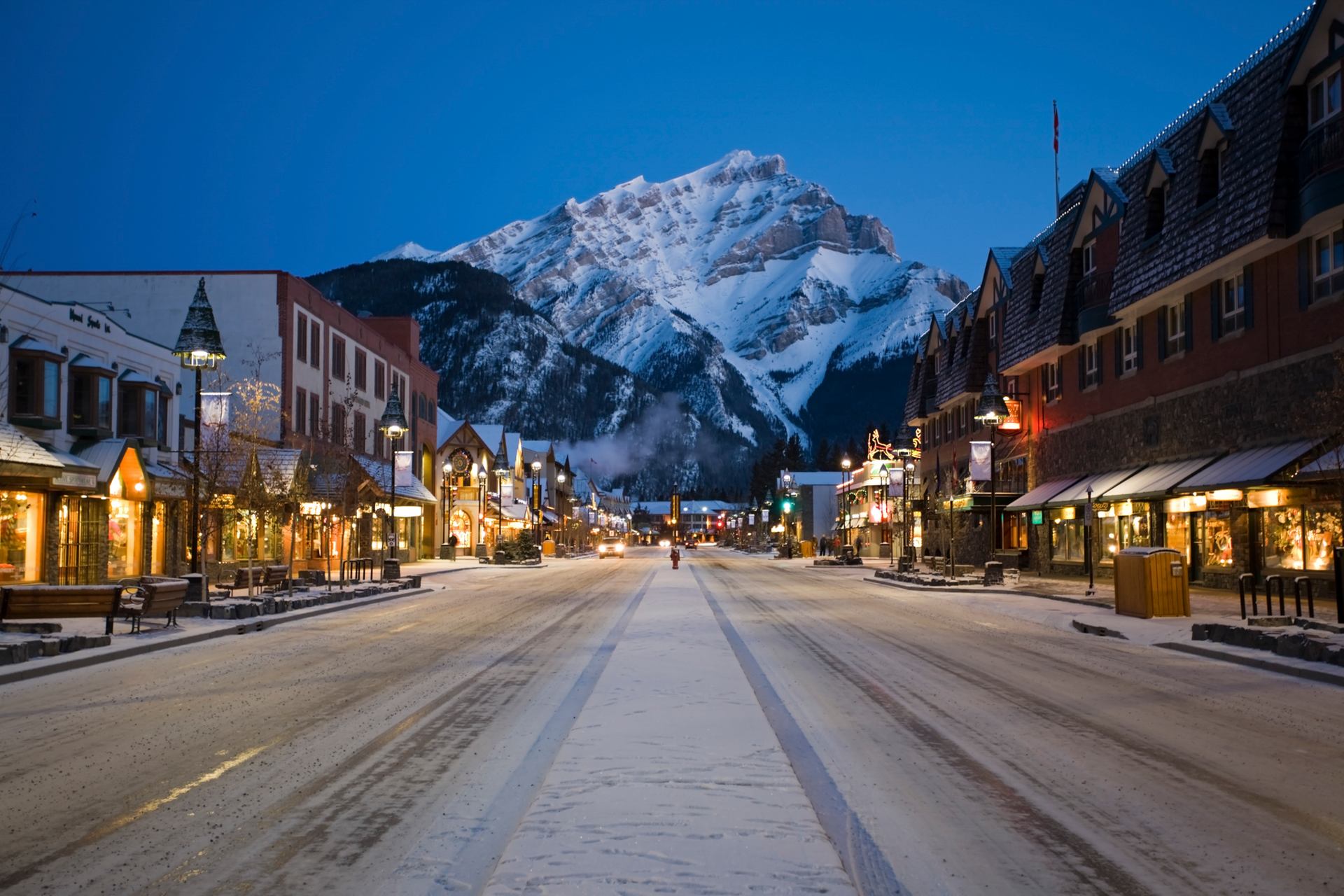 Snowy Banff Avenue lined with shops and lights, framed by majestic mountains at dusk.