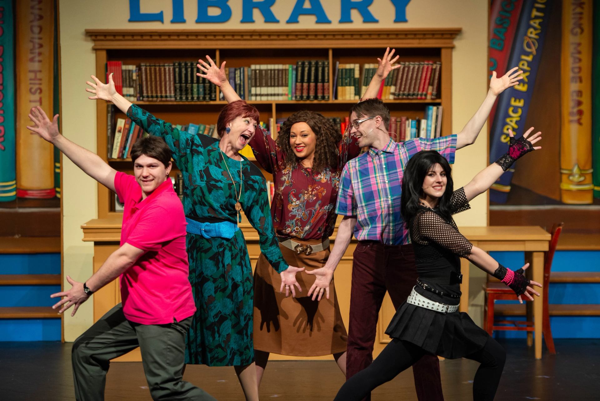 Stage performers pose energetically in colourful costumes in front of a library bookshelf backdrop.
