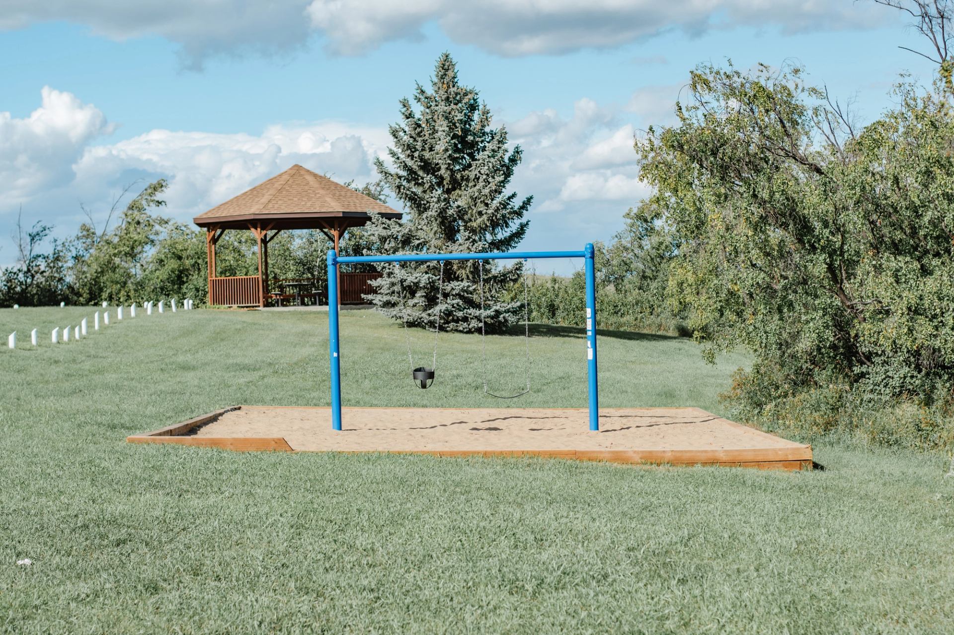 Empty blue swing set on sand beside gazebo and trees in grassy park.