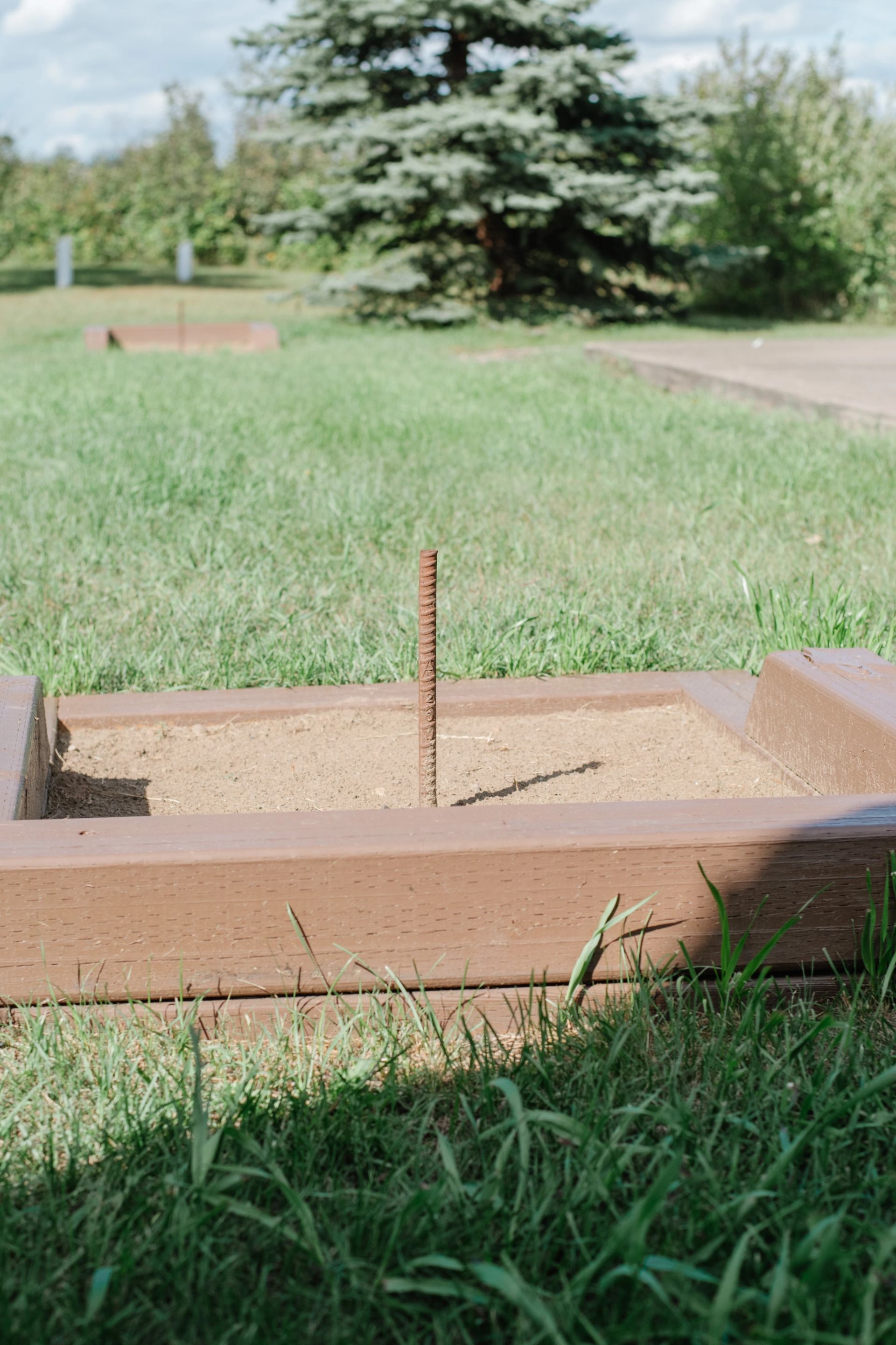 Horseshoe pit with metal stake in sand, surrounded by grass and trees.