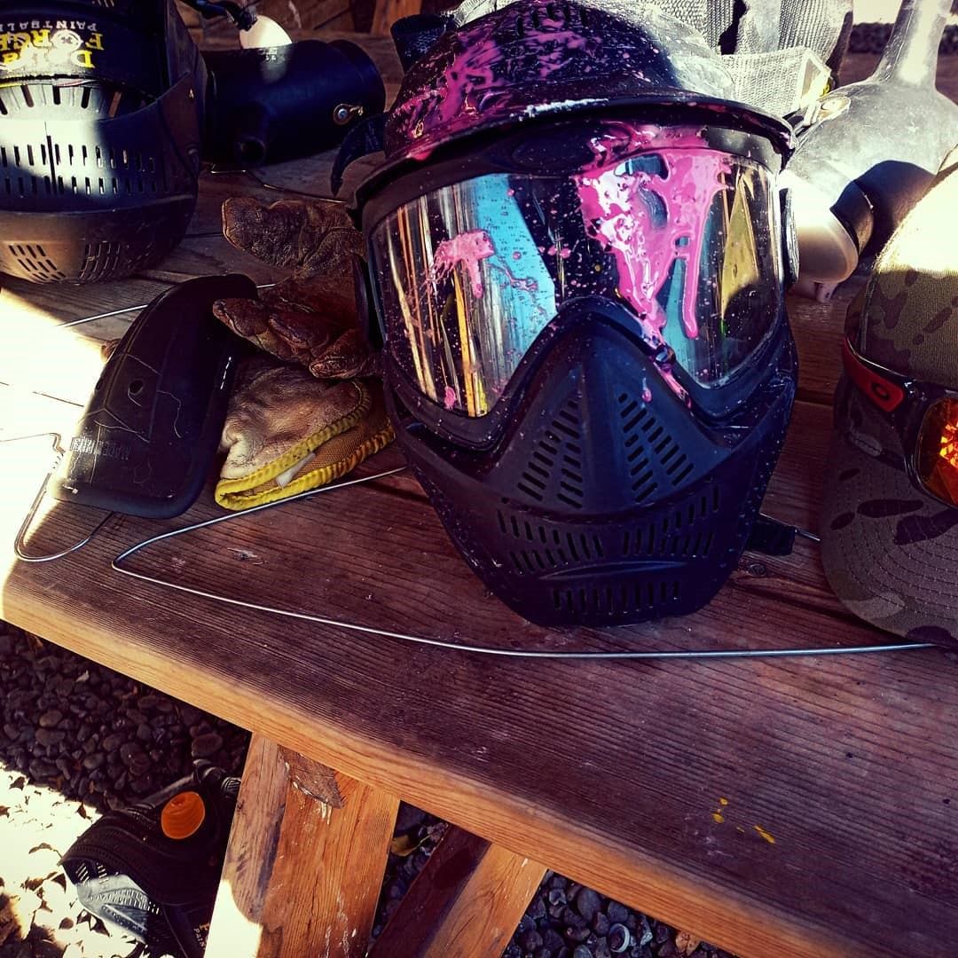 Paintball mask with pink splatter on visor beside gloves on wooden table.