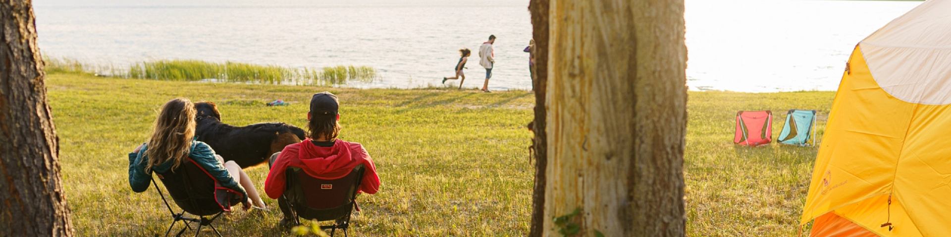 People sit in camp chairs by a lake while others walk and run near shoreline.