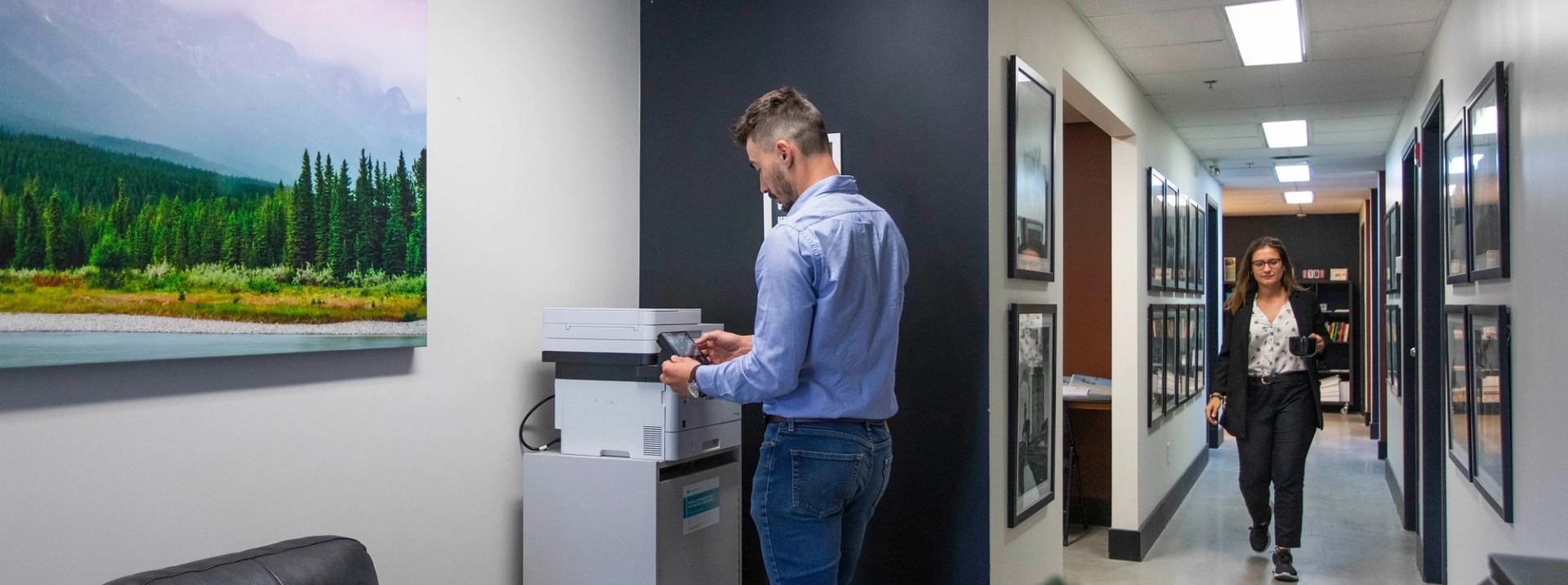 Two people working in a modern office hallway with framed photos and a printer station.