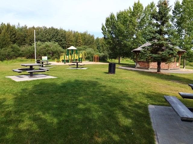 Park with picnic tables, playground, gazebo, and trees on green grass.