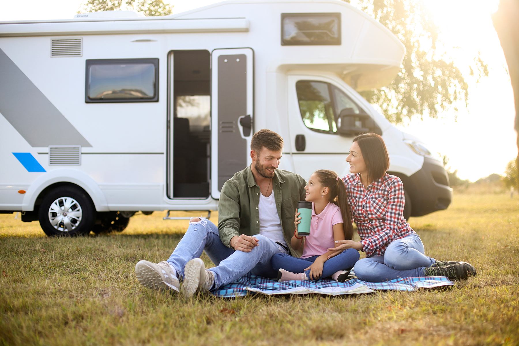 Group relaxing on a blanket near an RV parked on grass in a peaceful outdoor setting.