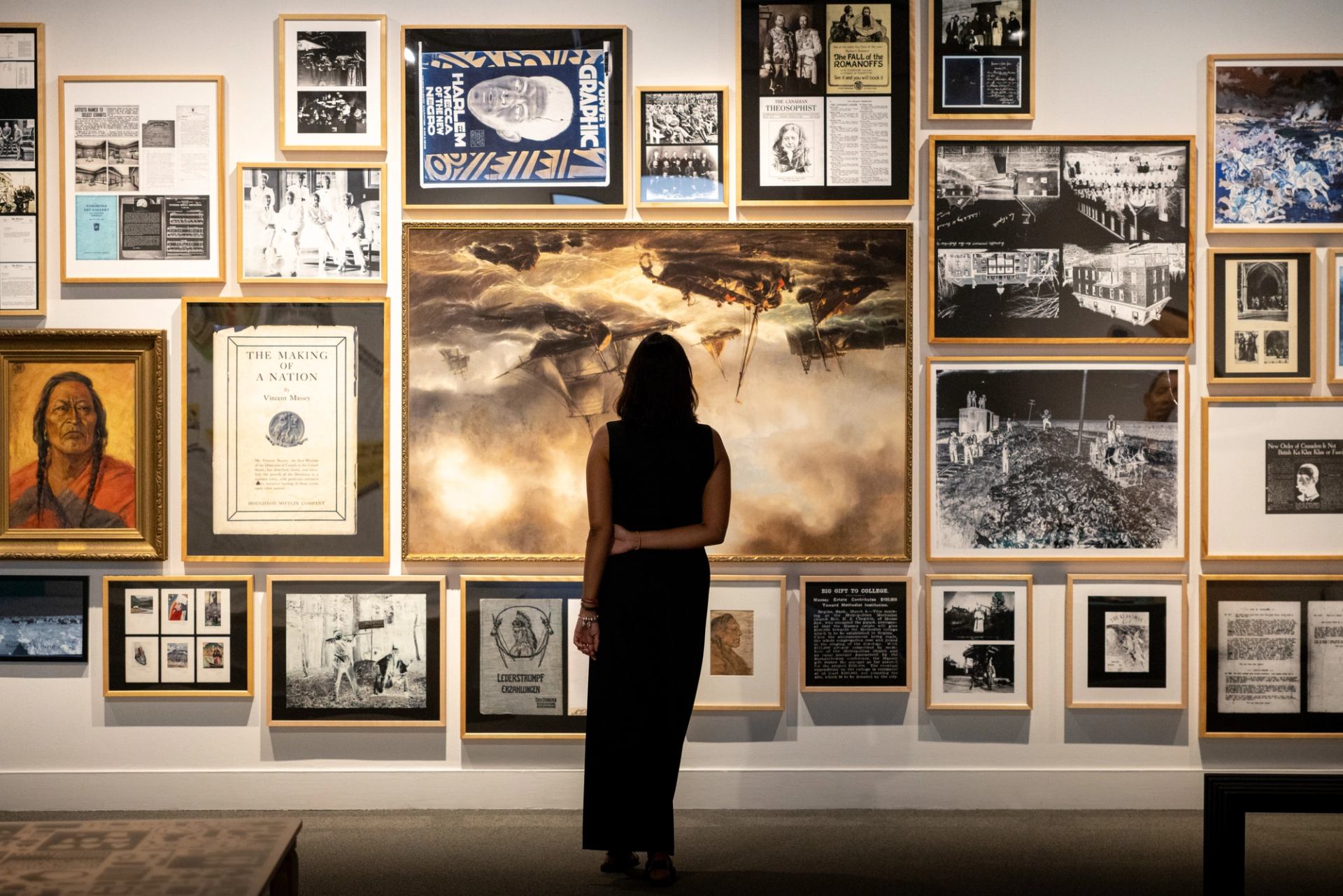Person in dark clothing stands before gallery wall of framed photos and paintings.