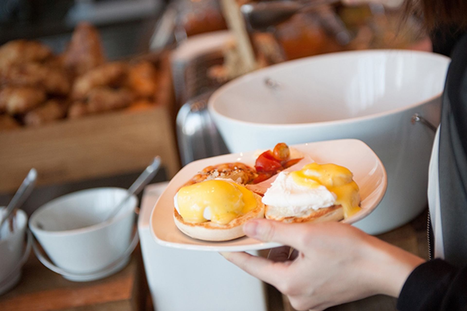A plate of eggs Benedict topped with hollandaise sauce is held near a buffet area with coffee cups and serving dishes in the background. The dish highlights a warm, freshly prepared breakfast offering.
