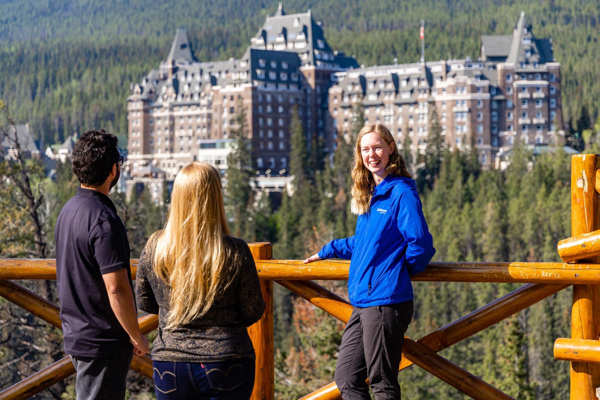 Three people stand at a wooden lookout facing a large forested mountain hotel.