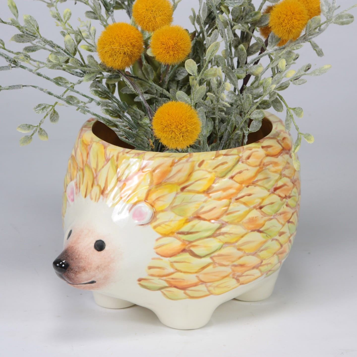 Glazed ceramic hedgehog planter with yellow pom-pom flowers on a white tabletop.