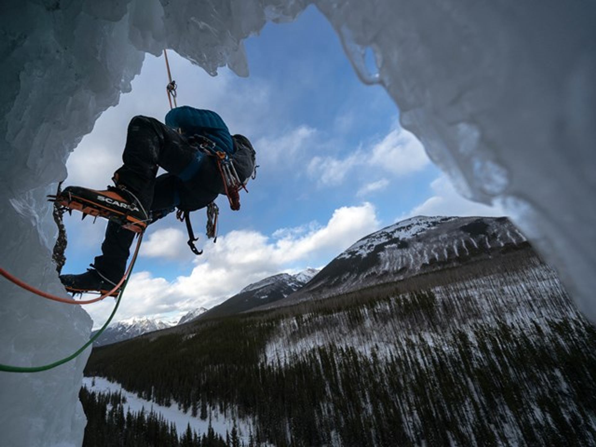 Ice climber suspended on a frozen formation overlooking snowy mountains and a forested valley.