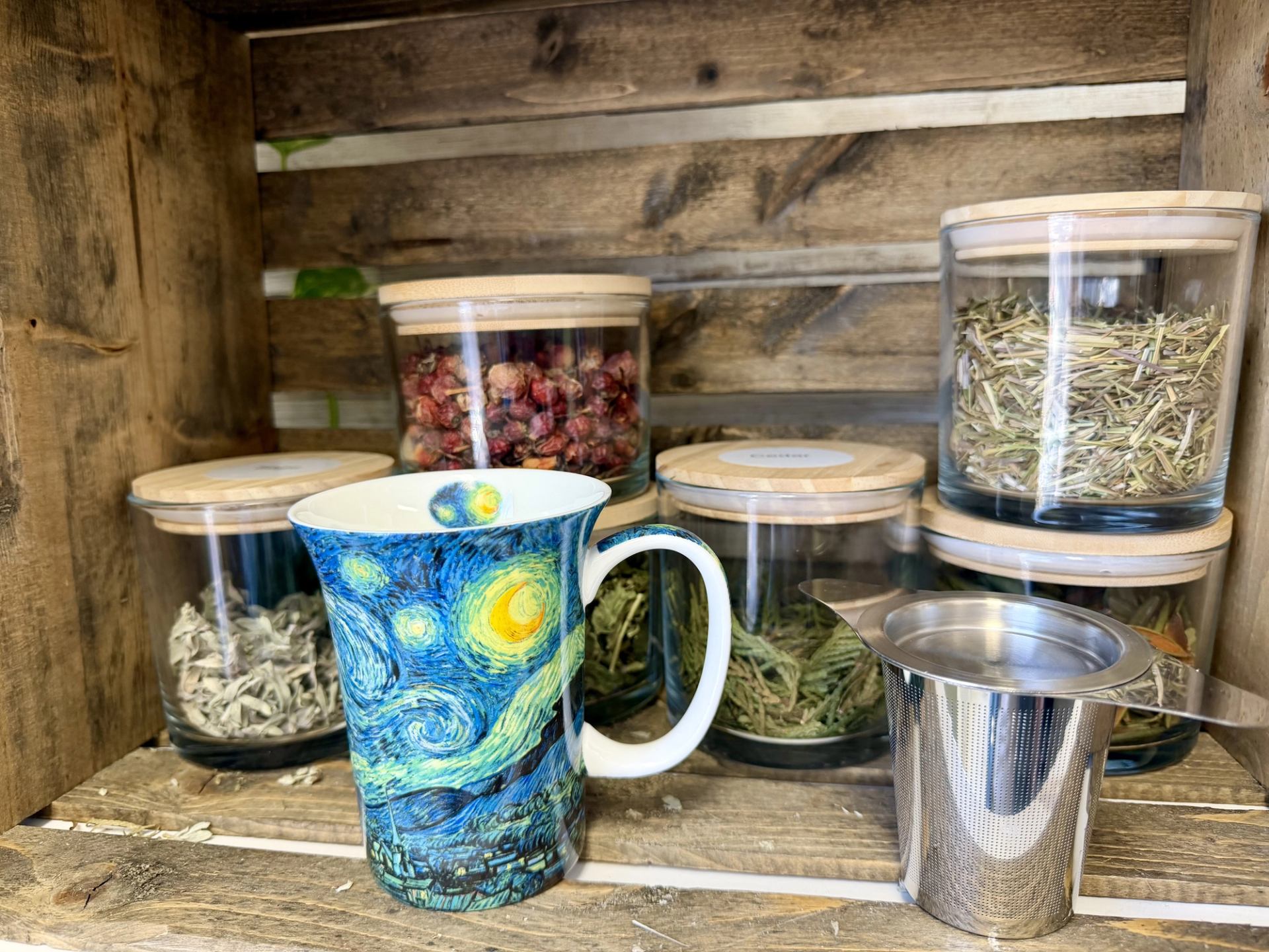 Jars of dried herbs sit on wooden shelves beside a mug and tea strainer.