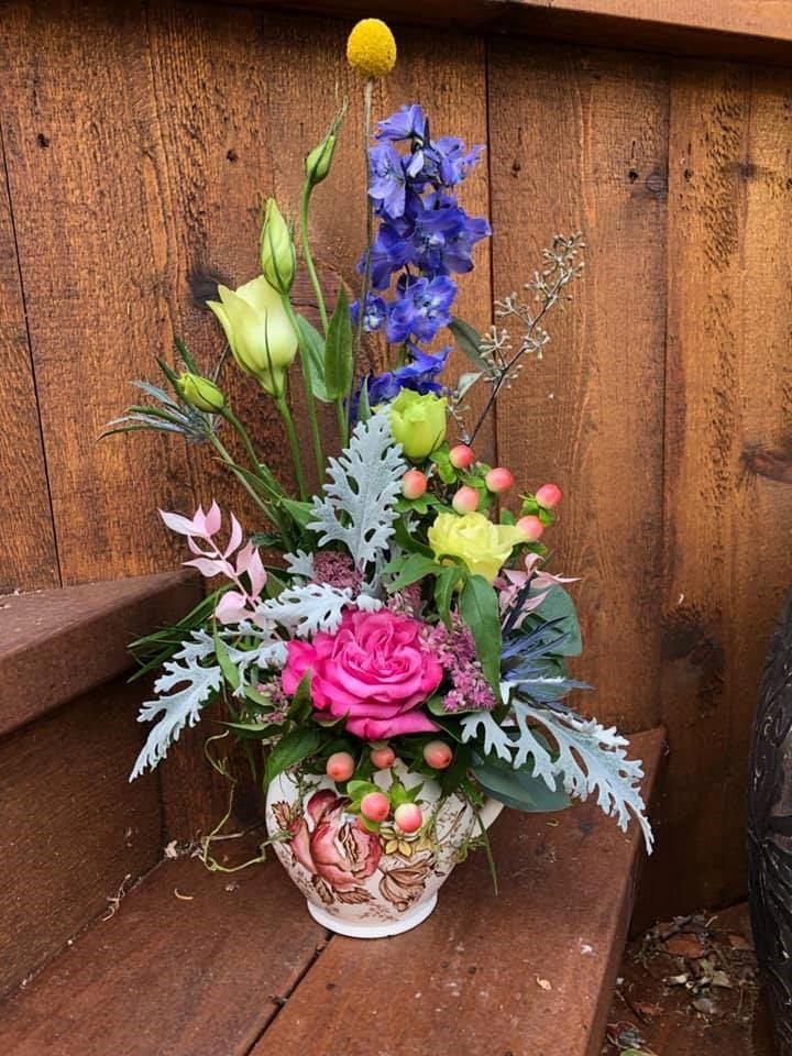 Floral arrangement in a decorative pot on a wooden bench beside a windowless wall.