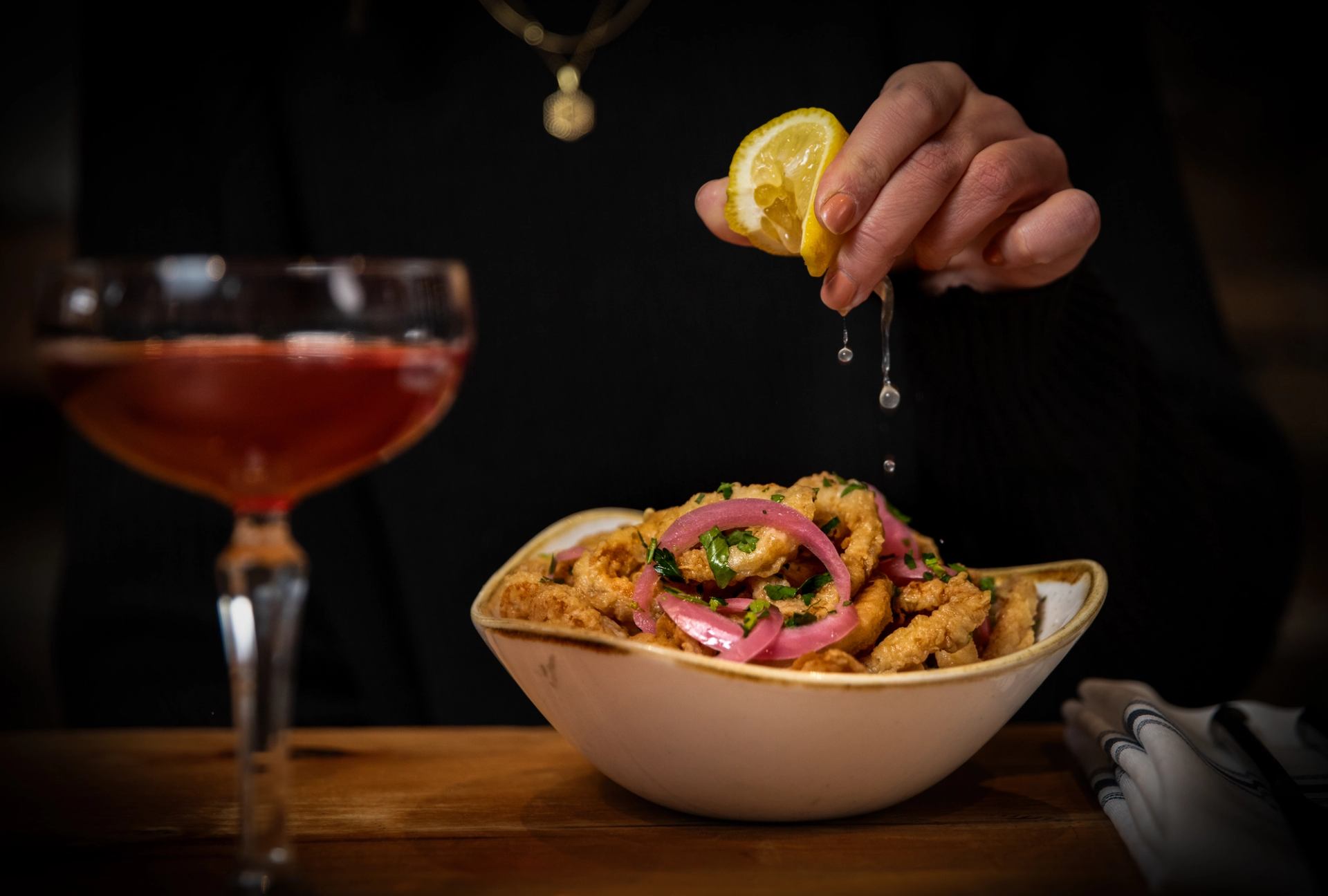Lemon being squeezed onto a bowl of fried kalamari with pickled red onions and tzatziki