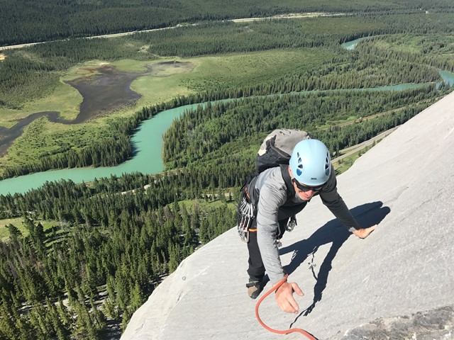 Climber ascending a steep rock face above a lush valley and winding river.