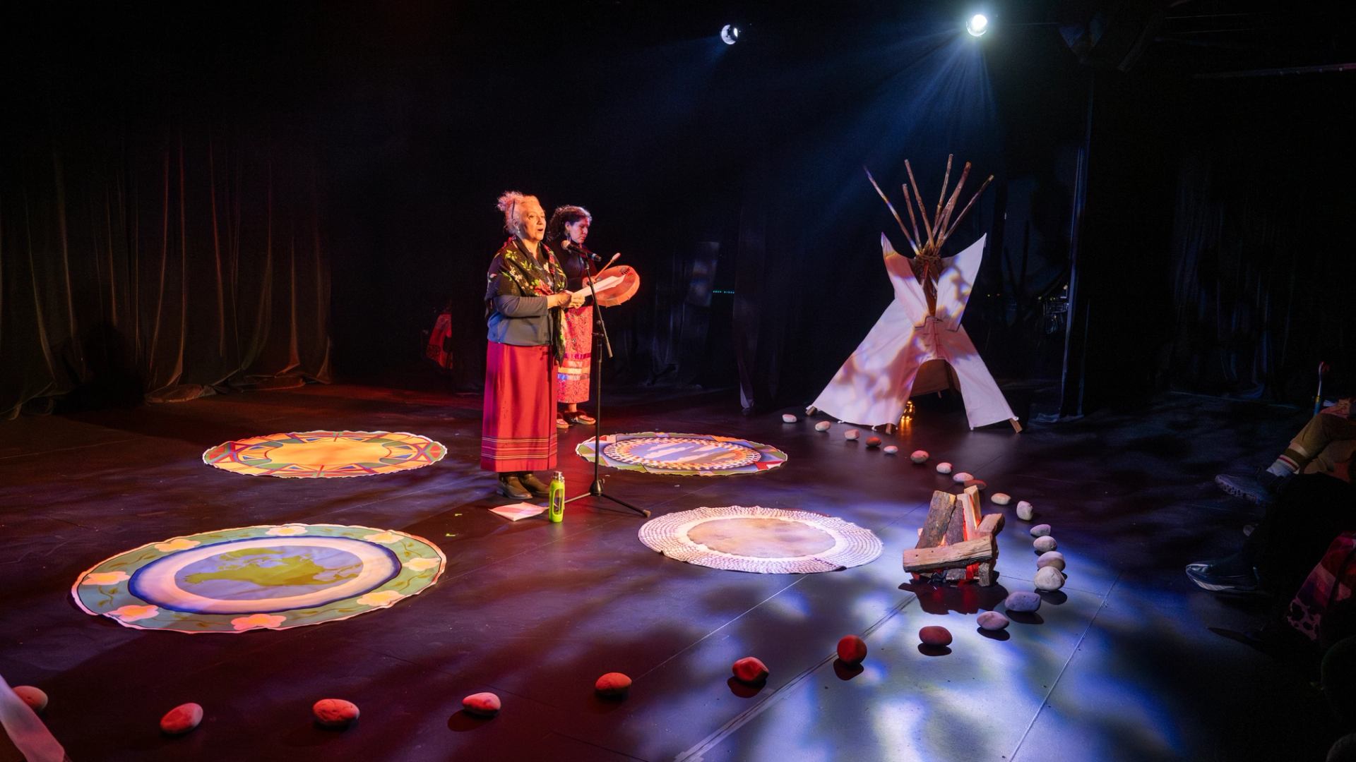 Two performers drumming onstage among circular artwork and a small tipi display.