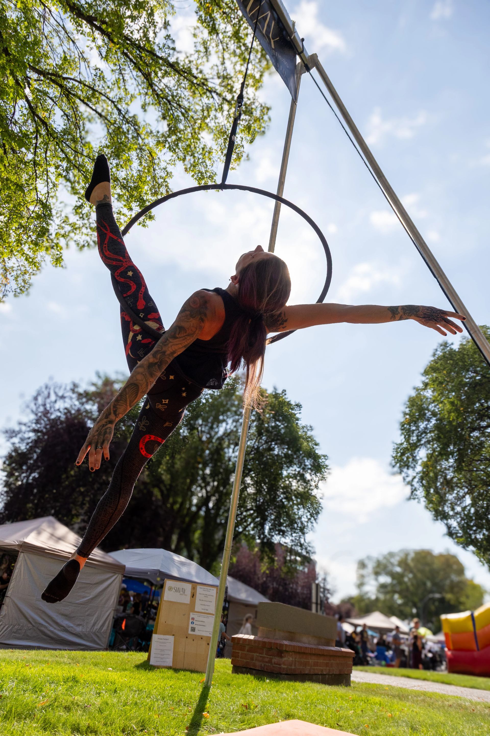Aerial performer stretches on suspended hoop above grass at outdoor fair with tents.