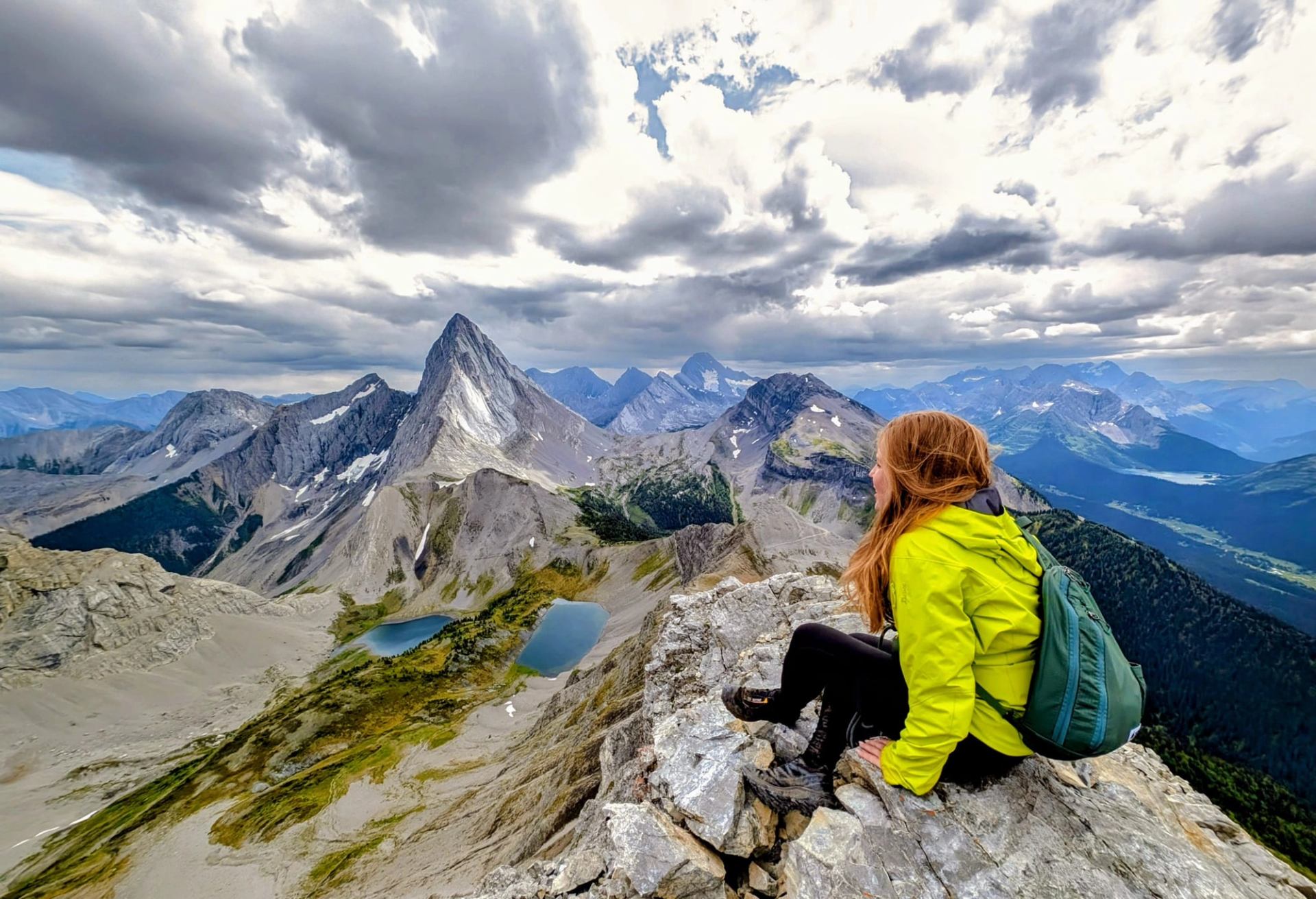 Hiker in green jacket sits on rocky ledge overlooking peaks and alpine lakes.