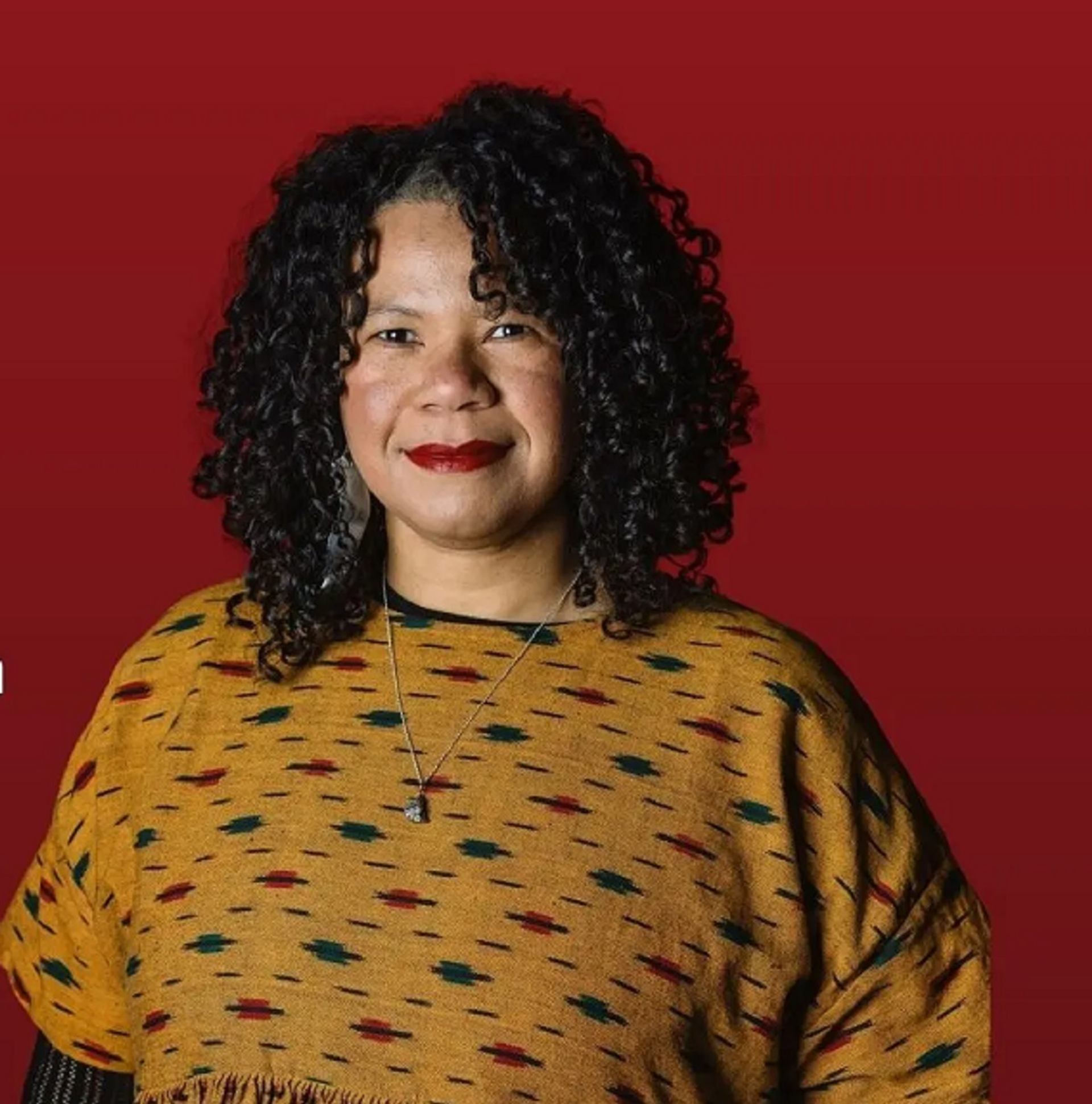 Person with curly hair wearing patterned mustard top stands against solid red background.