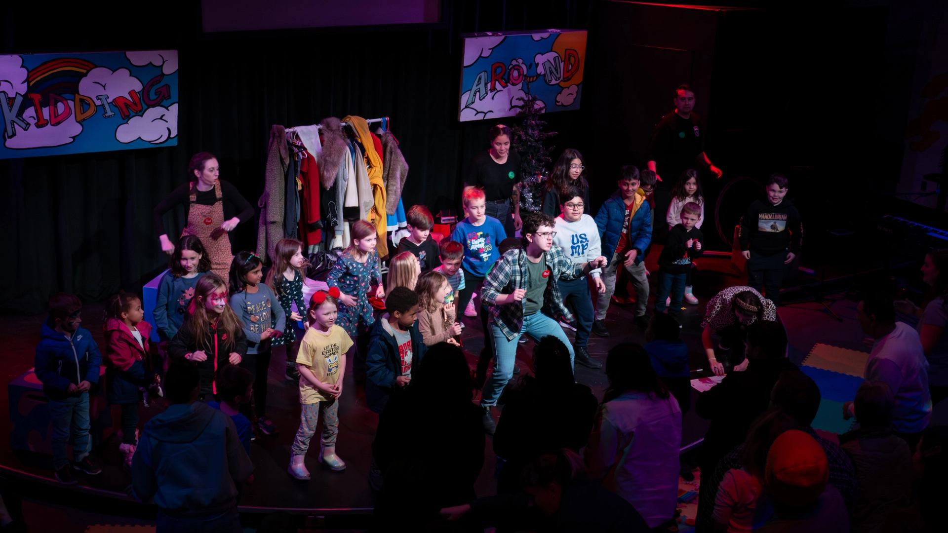 Children and adults gather on a small theatre stage, dancing under colourful lights.