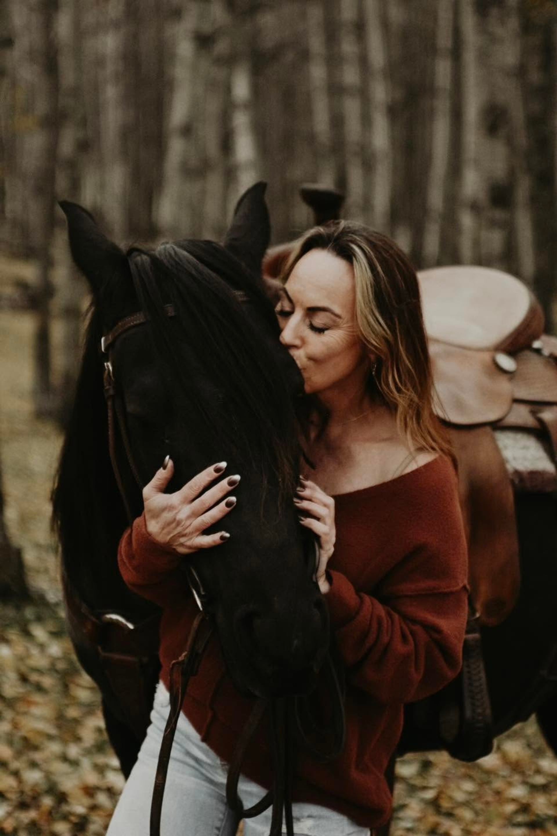 Person embraces and kisses a saddled black horse in a wooded area.