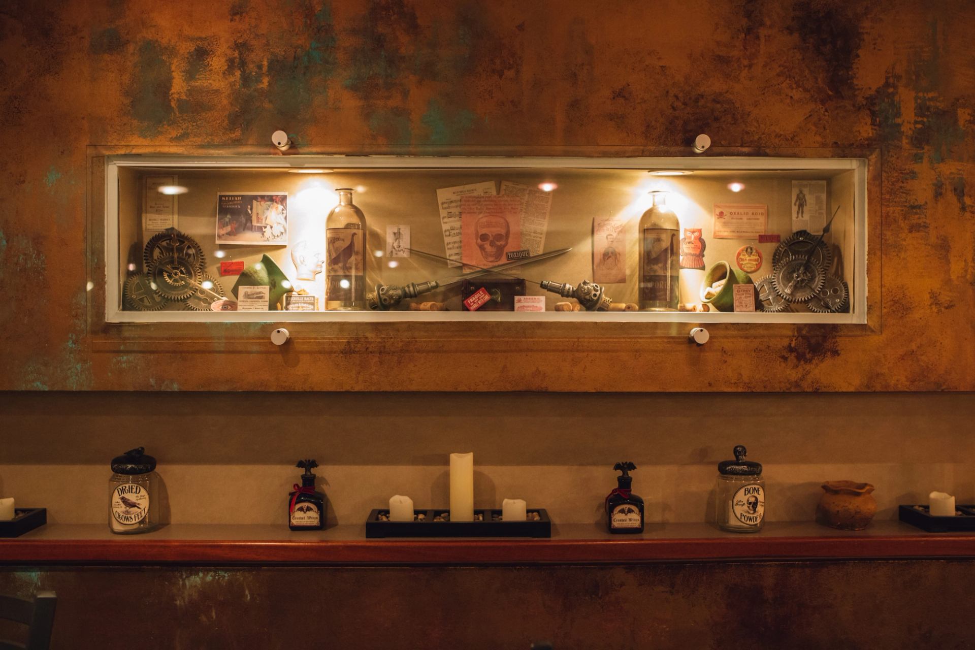 Illuminated display case with vintage bottles, gears, skull artwork and small labelled curiosities.
