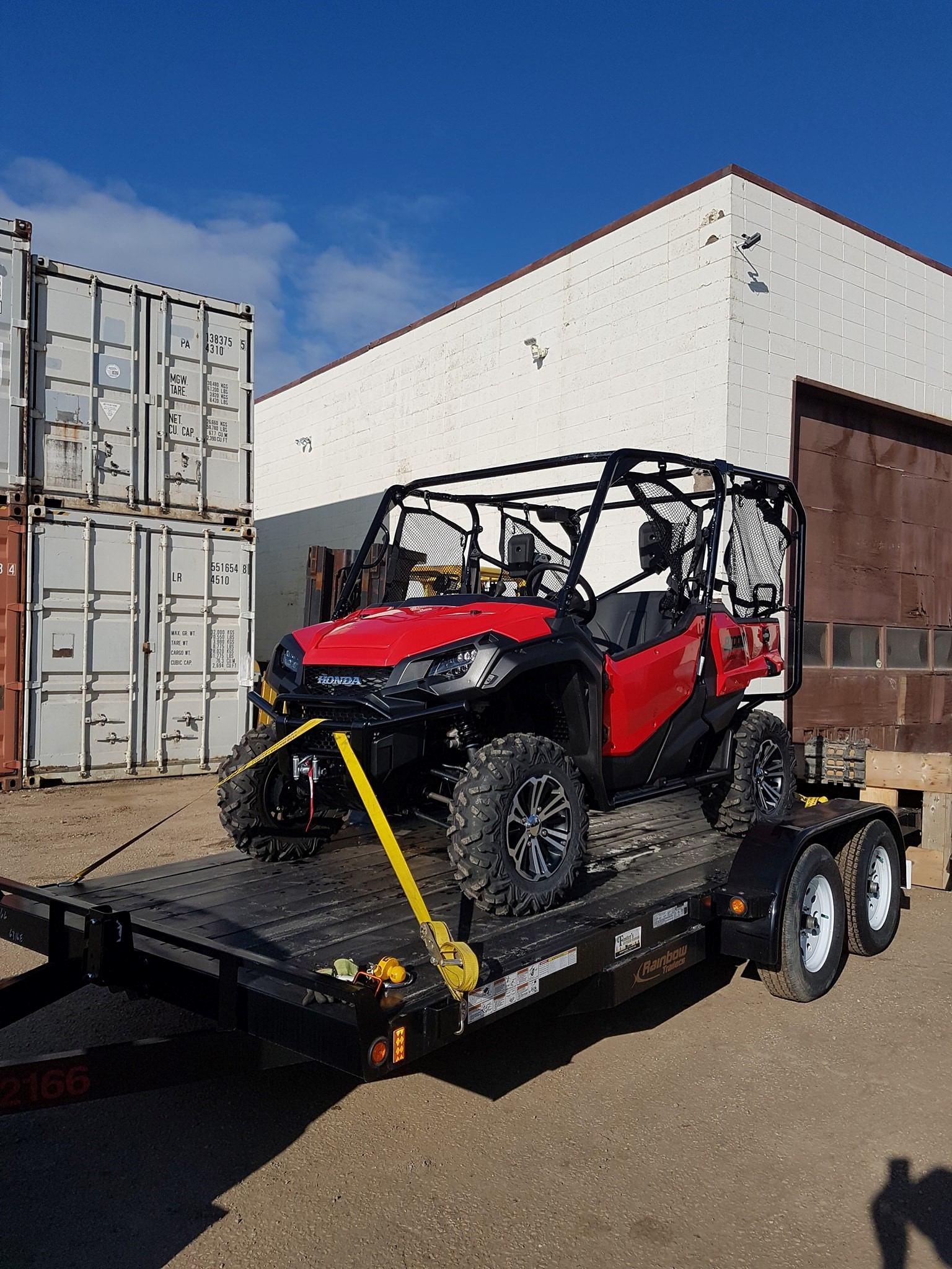Red and black off-road vehicle on trailer in industrial area with containers nearby.
