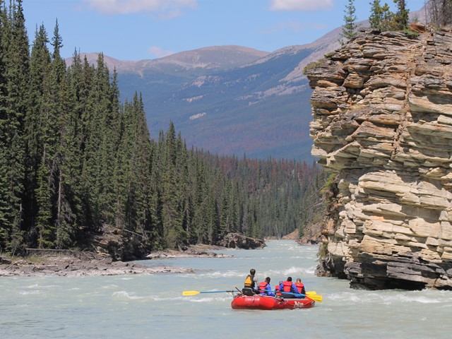 River rafting group in red inflatable raft on river beside rocky cliff and forest trees.