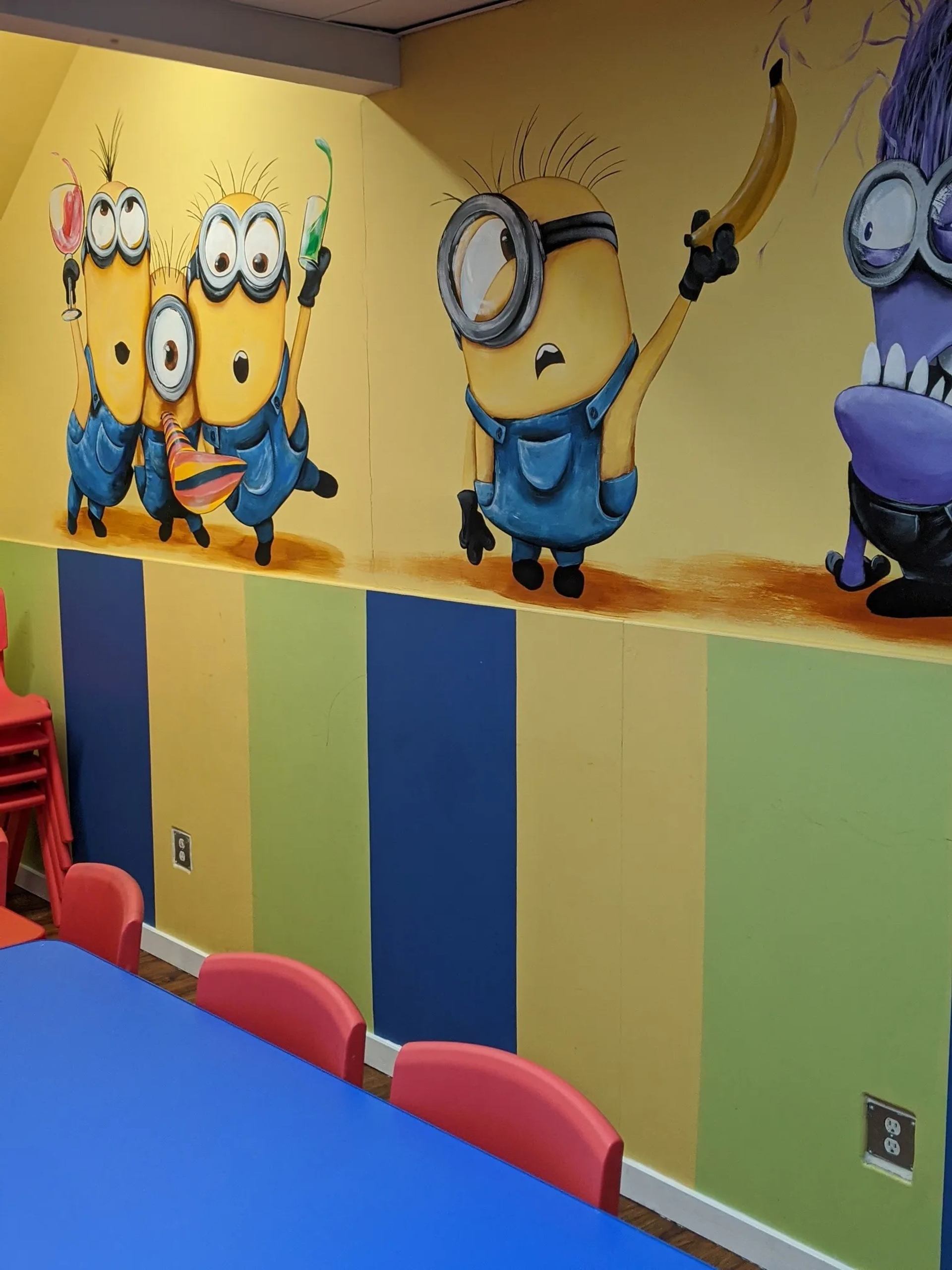 Minions wall mural above striped wall with blue table and red plastic chairs.