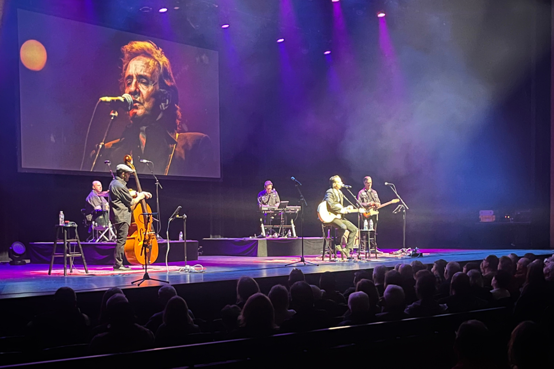 Band performs on stage under purple lights with singer projected on large screen.