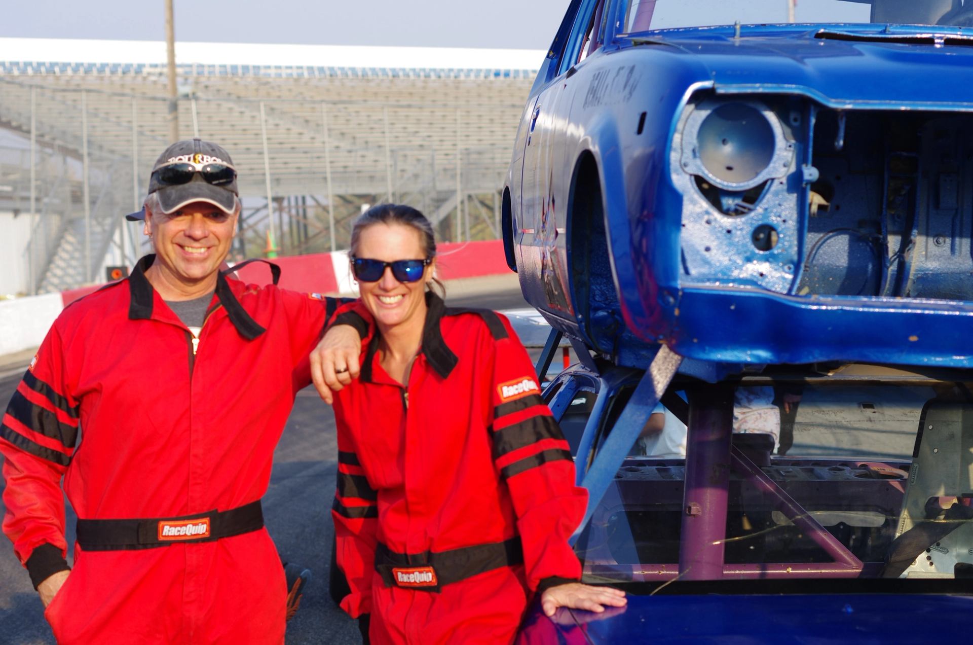 Two drivers in red racing suits pose next to a blue car at the track.