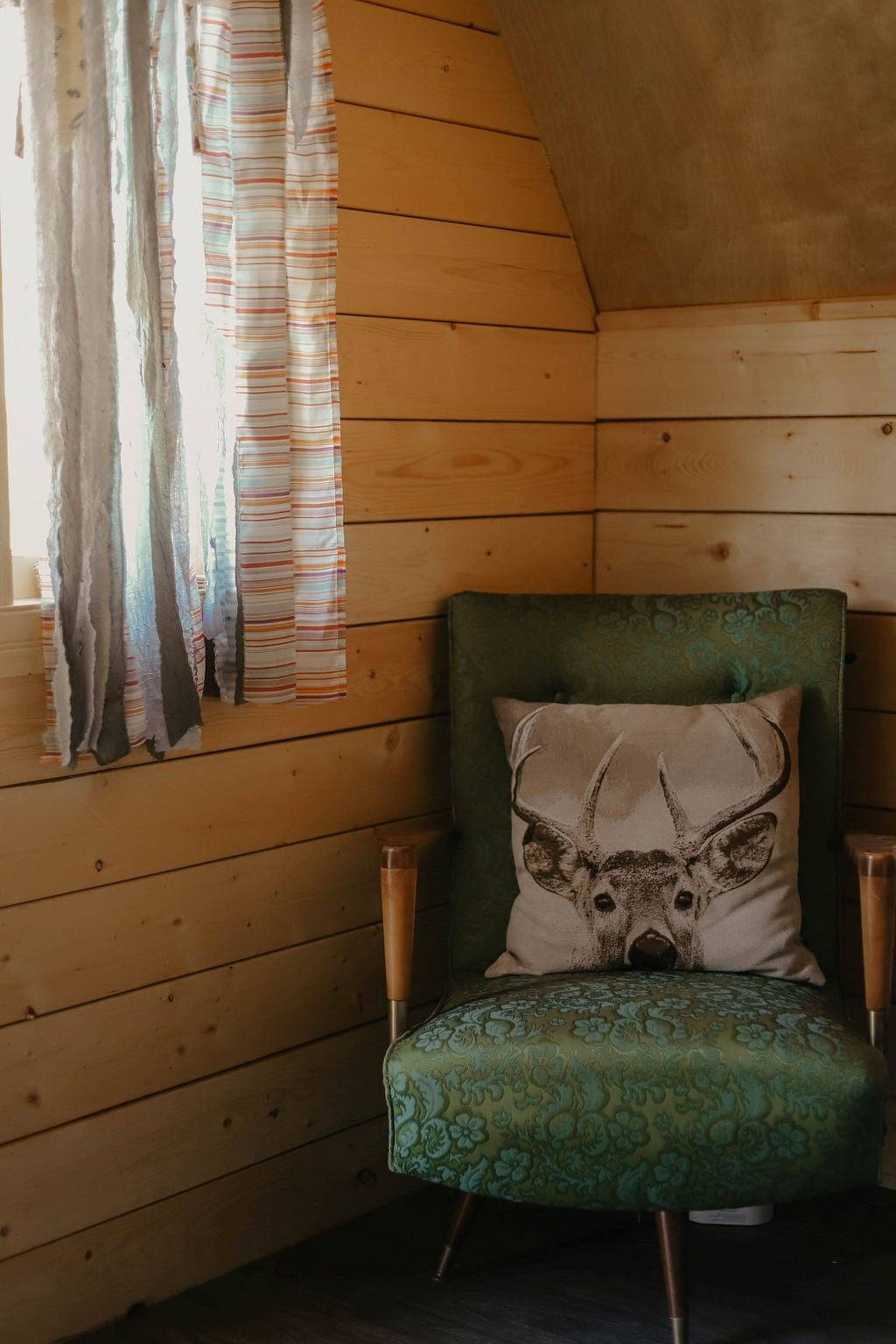 Cozy wooden room with a green armchair, featuring a decorative pillow with a deer print. Sunlight filters through striped curtains, creating a warm atmosphere.