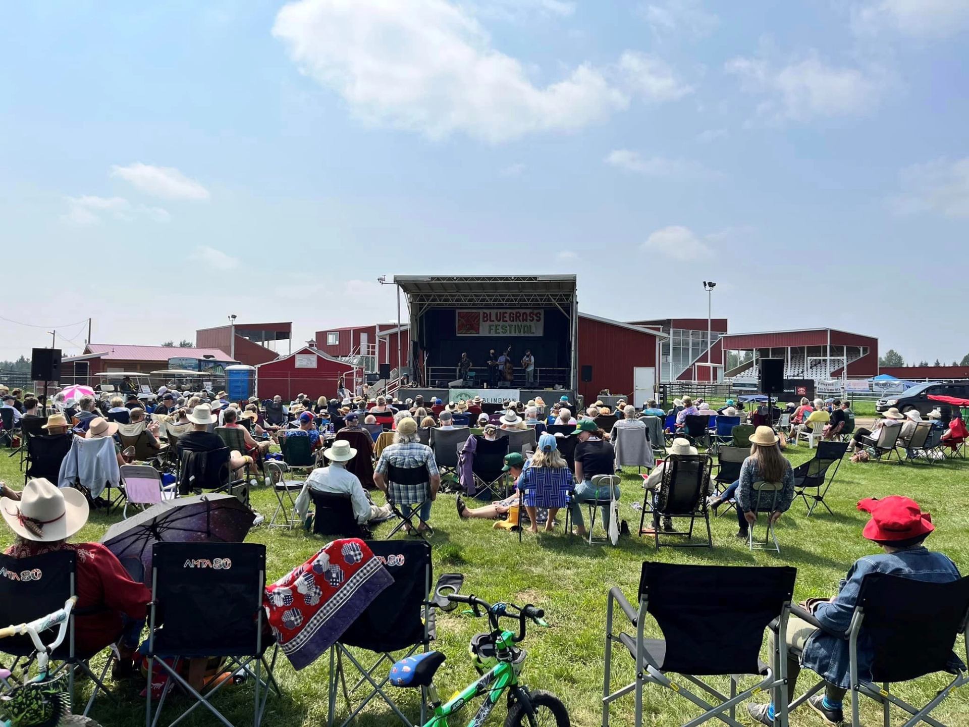 Outdoor bluegrass festival stage with band and audience seated in folding chairs.