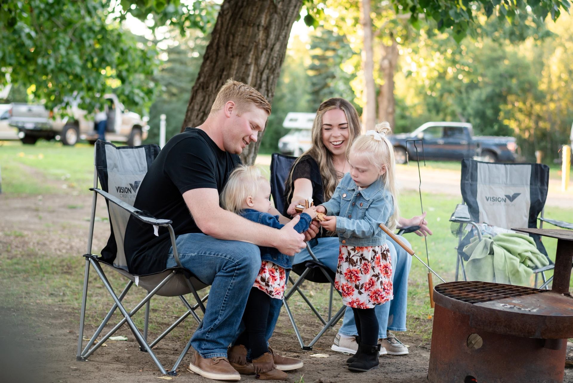Adults and children sit in camping chairs, roasting food over a metal firepit.