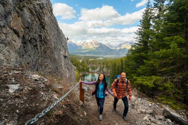 A couple walking up the Grassi Lakes Trail past the section where rock climbers climb in the distance.